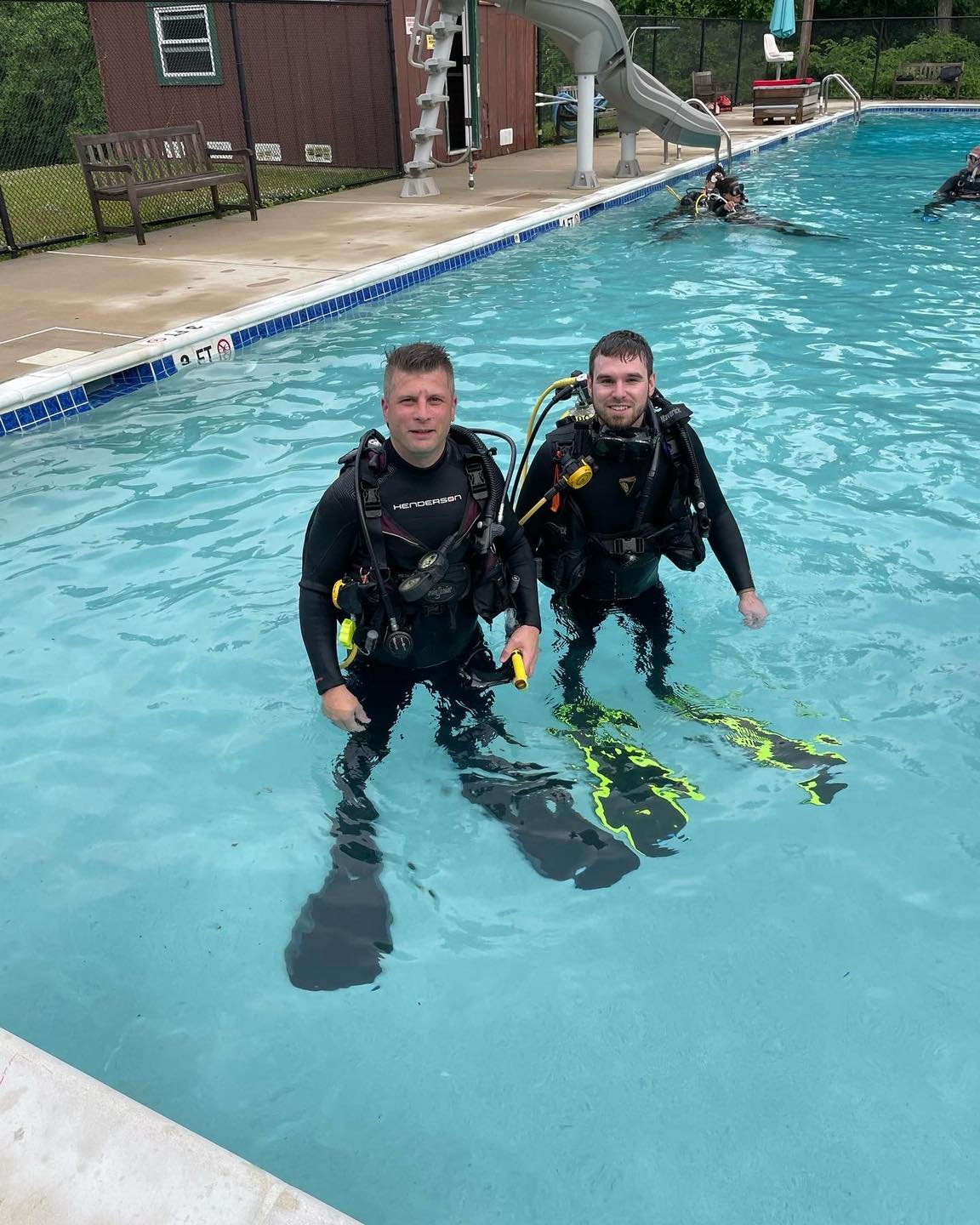 Two men in scuba gear standing in a swimming pool, smiling at the camera.