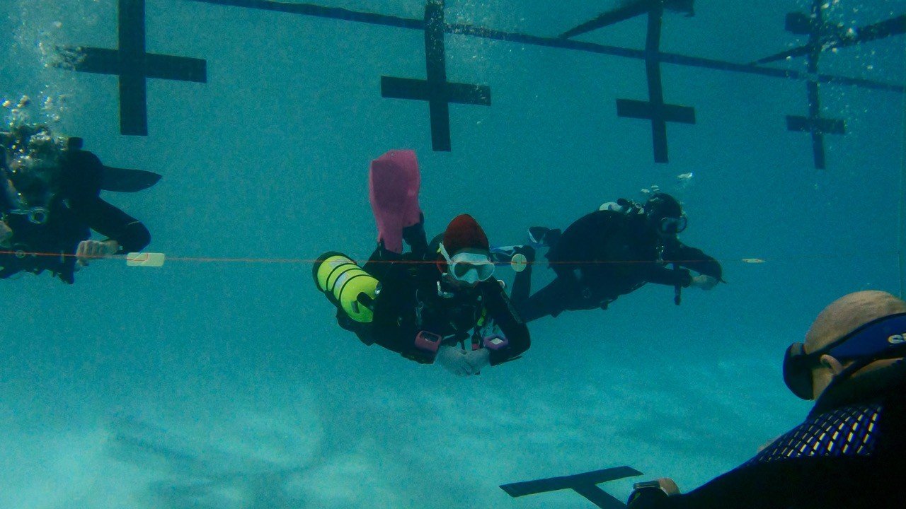 Scuba divers underwater near a pool's bottom, with safety barriers and crosses visible above.