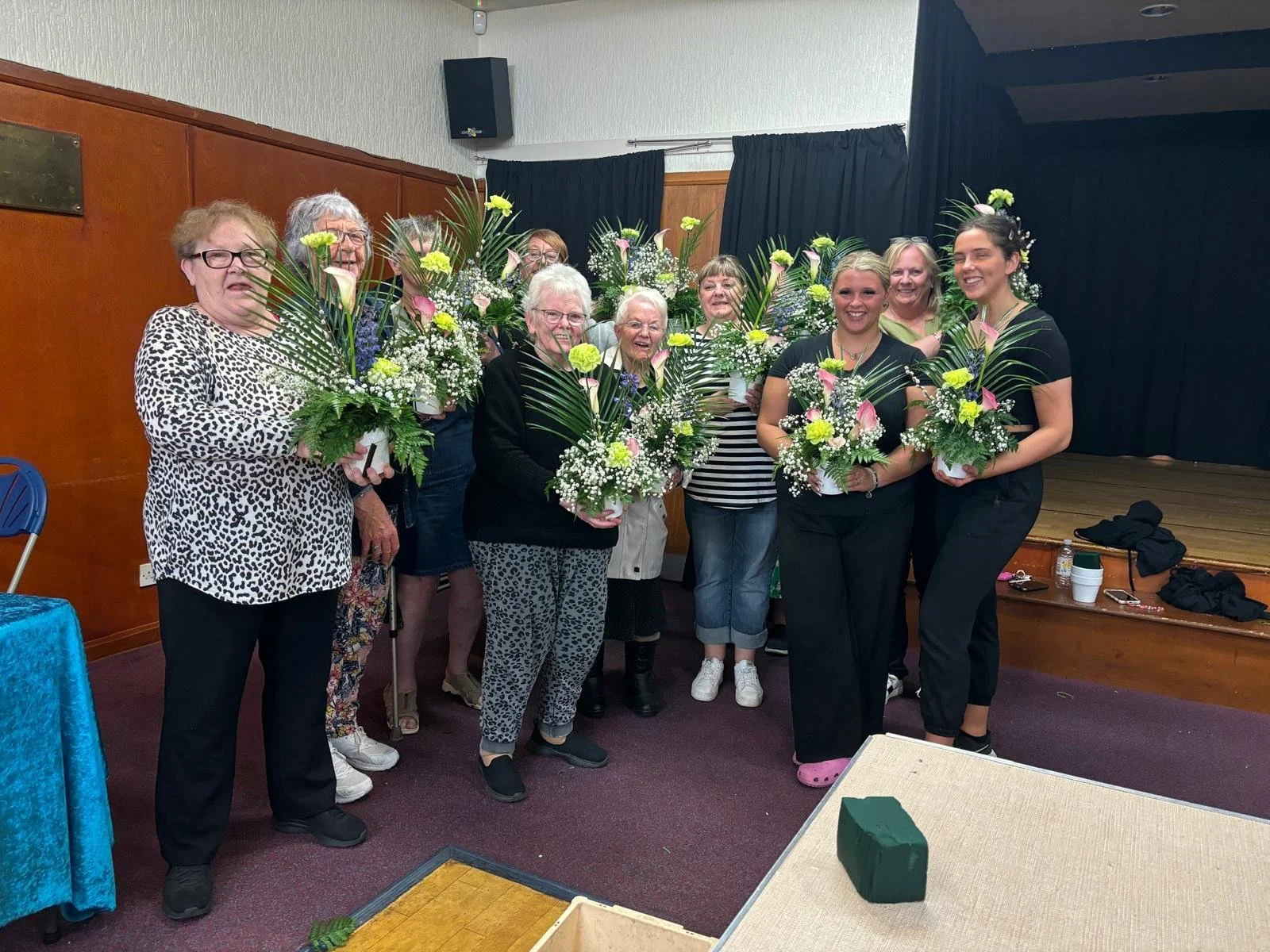 A group of nine women of various ages standing indoors, holding flower arrangements, smiling for a photo in front of a stage with black curtains.