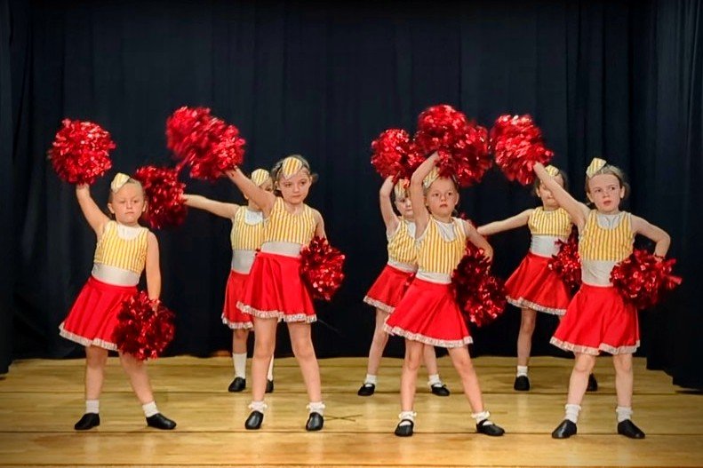 Young girls in yellow and red cheerleading costumes performing a cheer routine with pom-poms on a stage with a black curtain background.