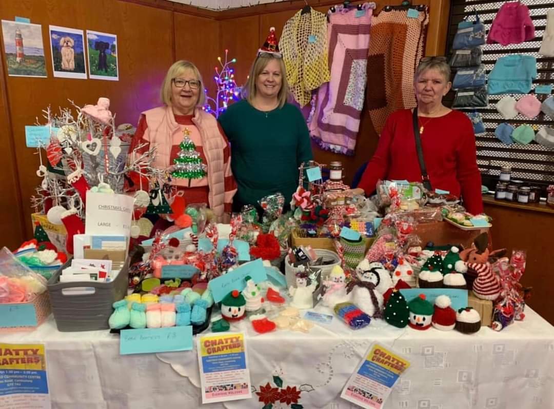 Three women standing behind a table filled with Christmas crafts and decorations at a craft fair.