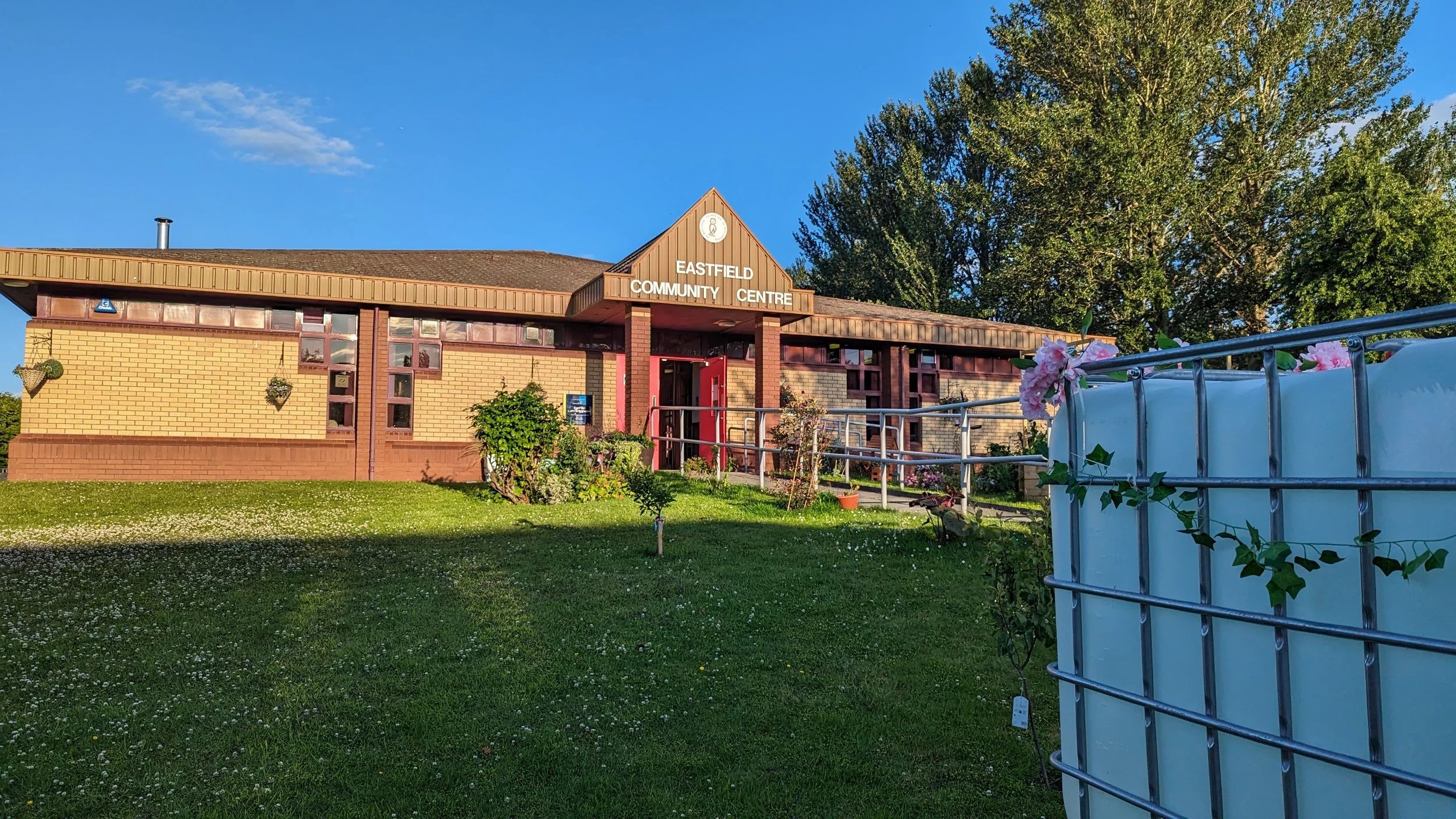Eastfield Community Centre building on a sunny day, with red double doors under a peaked roof sign, surrounded by well-kept grass, plants, and trees.