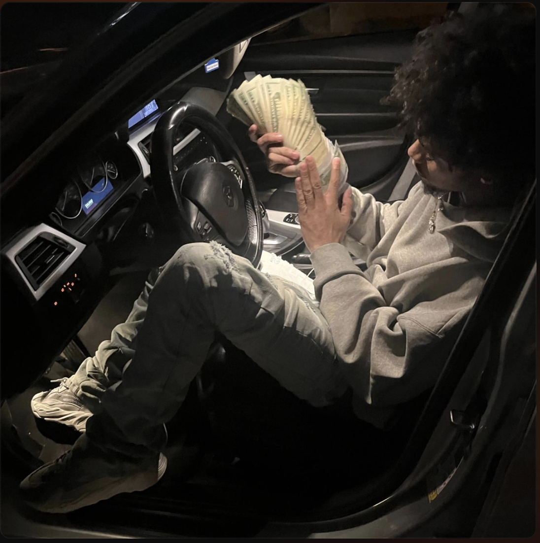 Person sitting in the driver's seat of a car, holding a large fan of US $100 bills, with cash in hand.