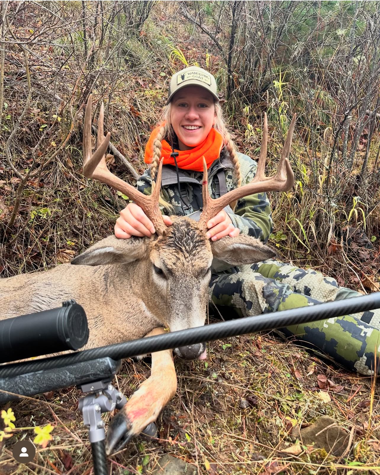 A woman in camouflage outdoor gear and an orange neck gaiter smiling and holding the antlers of a large deer lying on the ground in a wooded area.