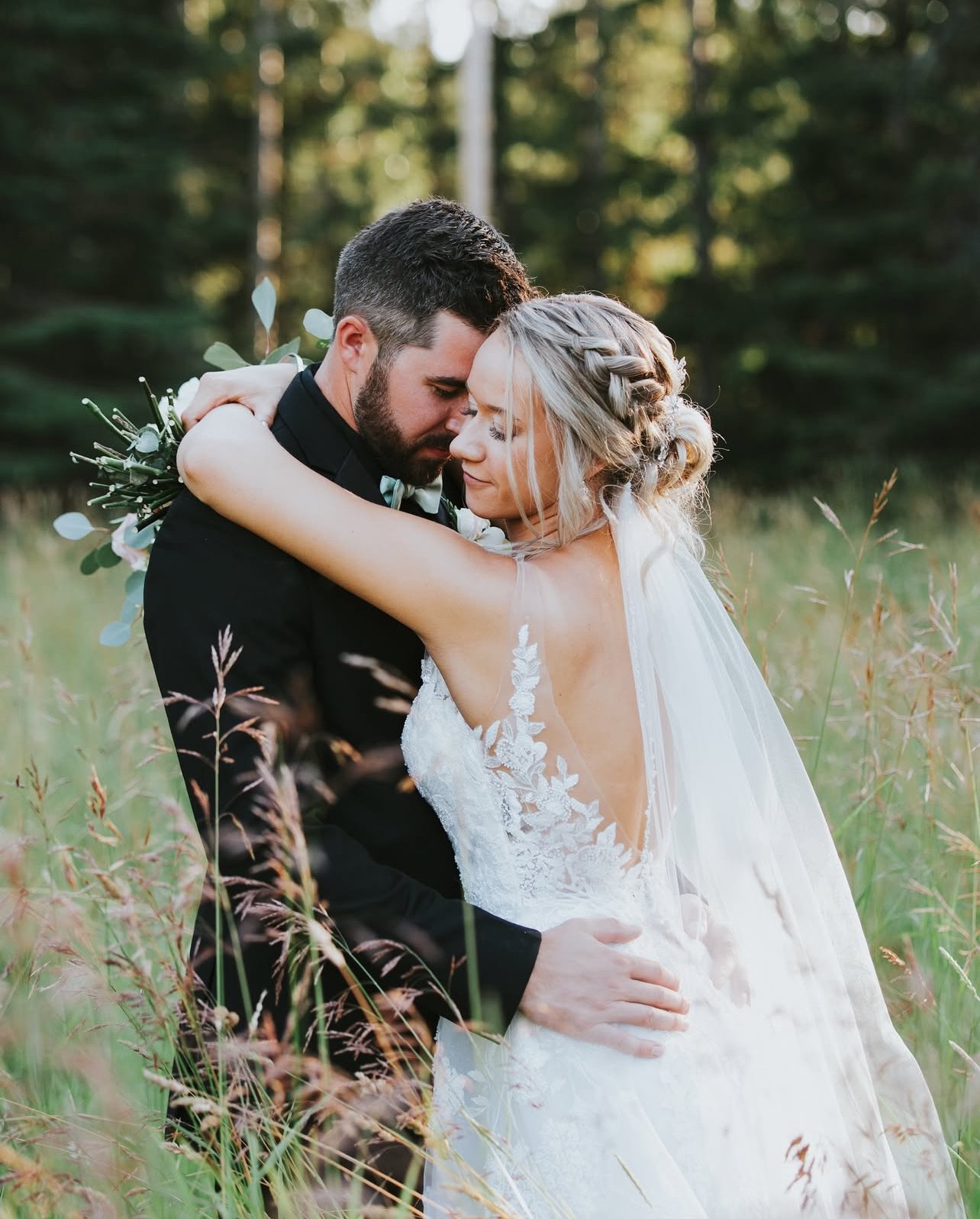 A bride and groom embracing outdoors in a field of tall grass, close with foreheads touching, surrounded by green trees.