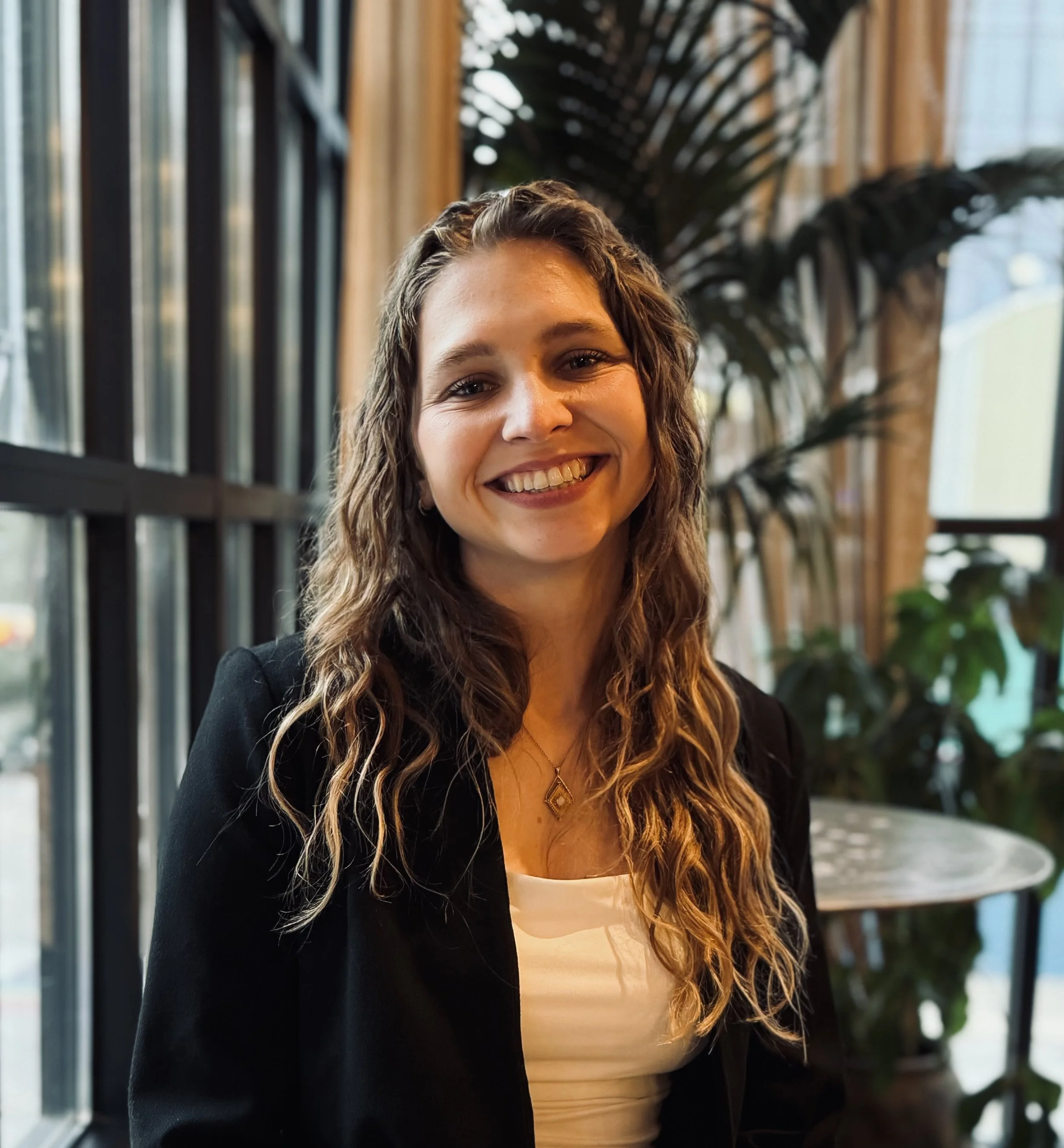 A woman with long wavy brown hair smiling, wearing a black blazer and a white top, sitting in a well-lit indoor space with large windows and lush green plants in the background.
