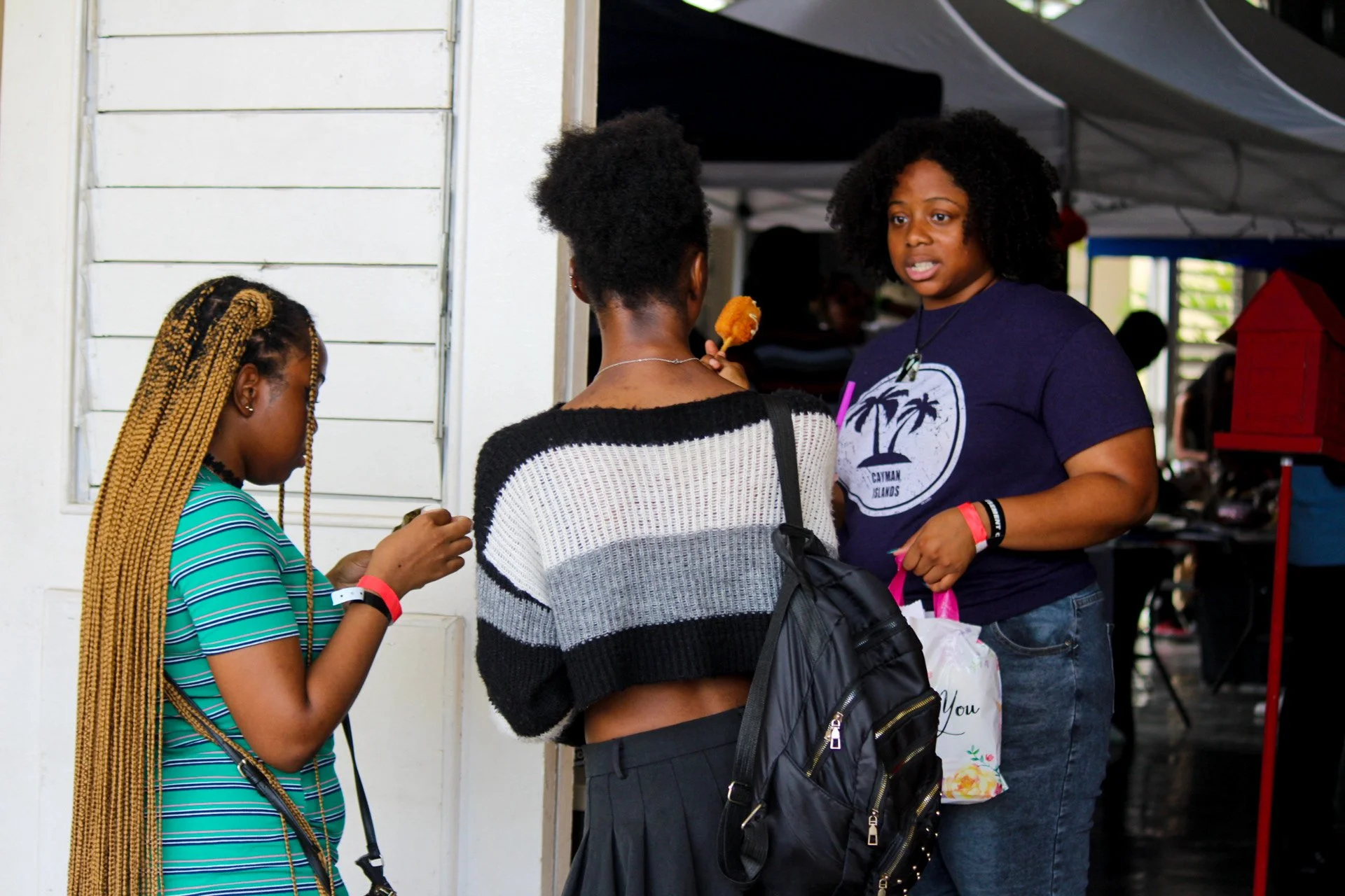 Three women having a conversation at an outdoor market or fair, one holding a fried food item, in front of tents and stalls.
