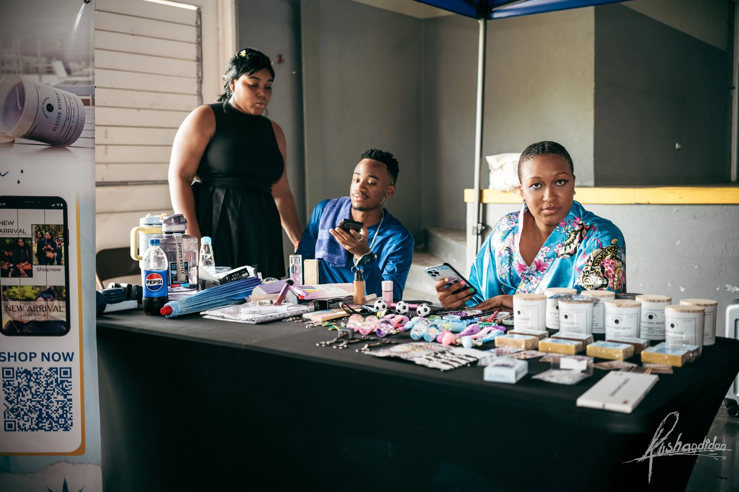 Three people sit behind a table displaying various products, with one woman standing, in an indoor setting. The table is covered with items like cosmetics, small toys, and packaged goods.