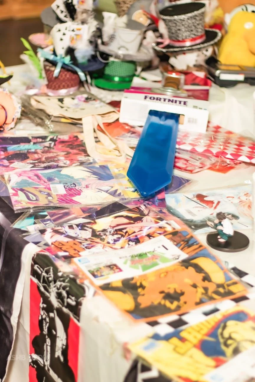 A cluttered table with various items including trading cards, a blue card holder, a Fortnite item box, small figurines, and hats. The background shows additional hats and miscellaneous items.