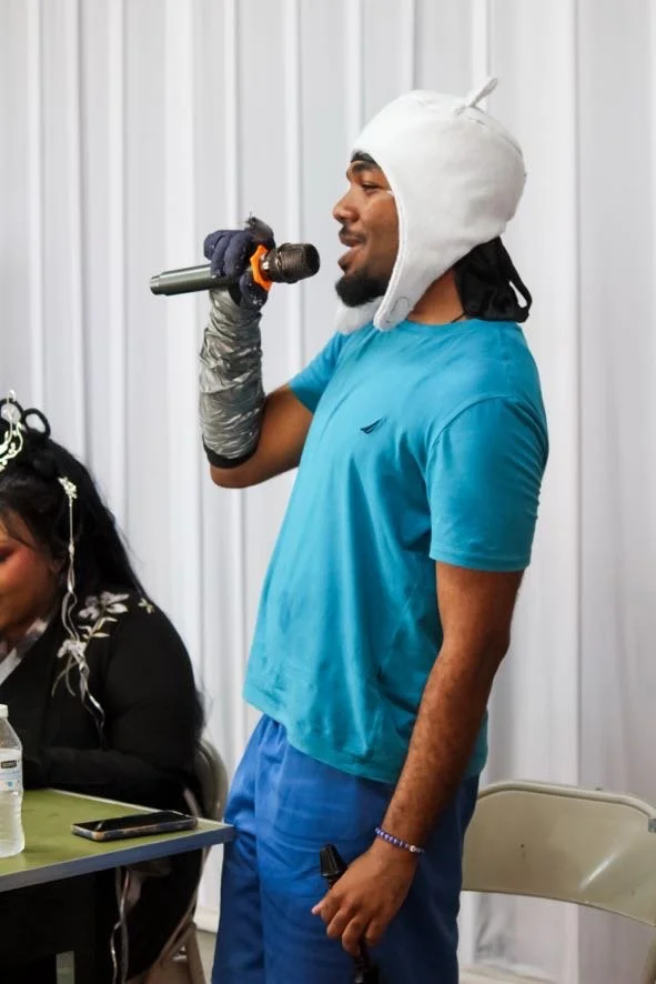 A man wearing a white hat, blue t-shirt, and gloves is holding a microphone, possibly speaking or singing, while standing in a room with white curtains.