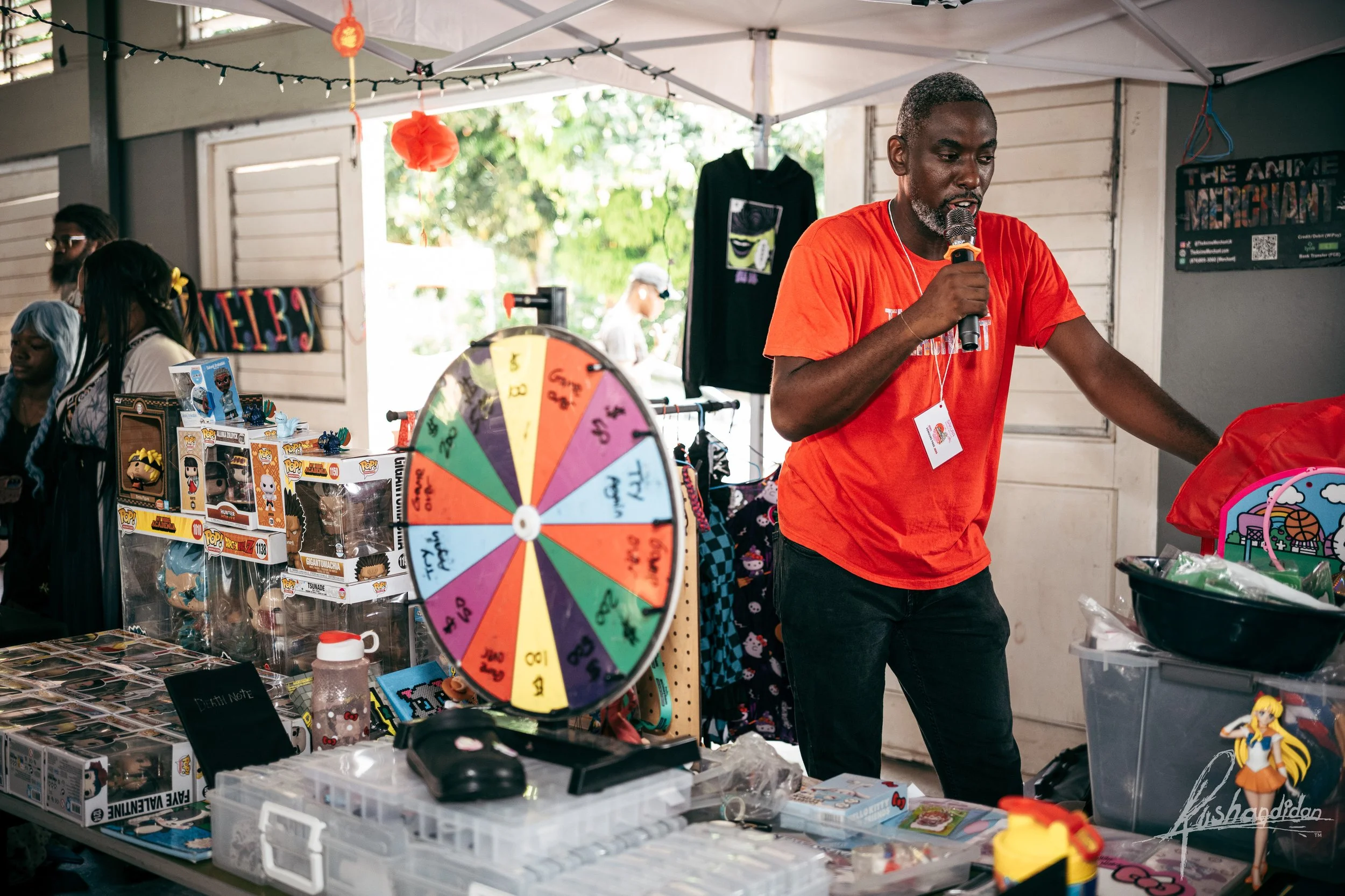 Man wearing a red shirt holding a microphone at a vendor booth with a colorful spinning prize wheel, toys, and merchandise, under a tent at an outdoor event.