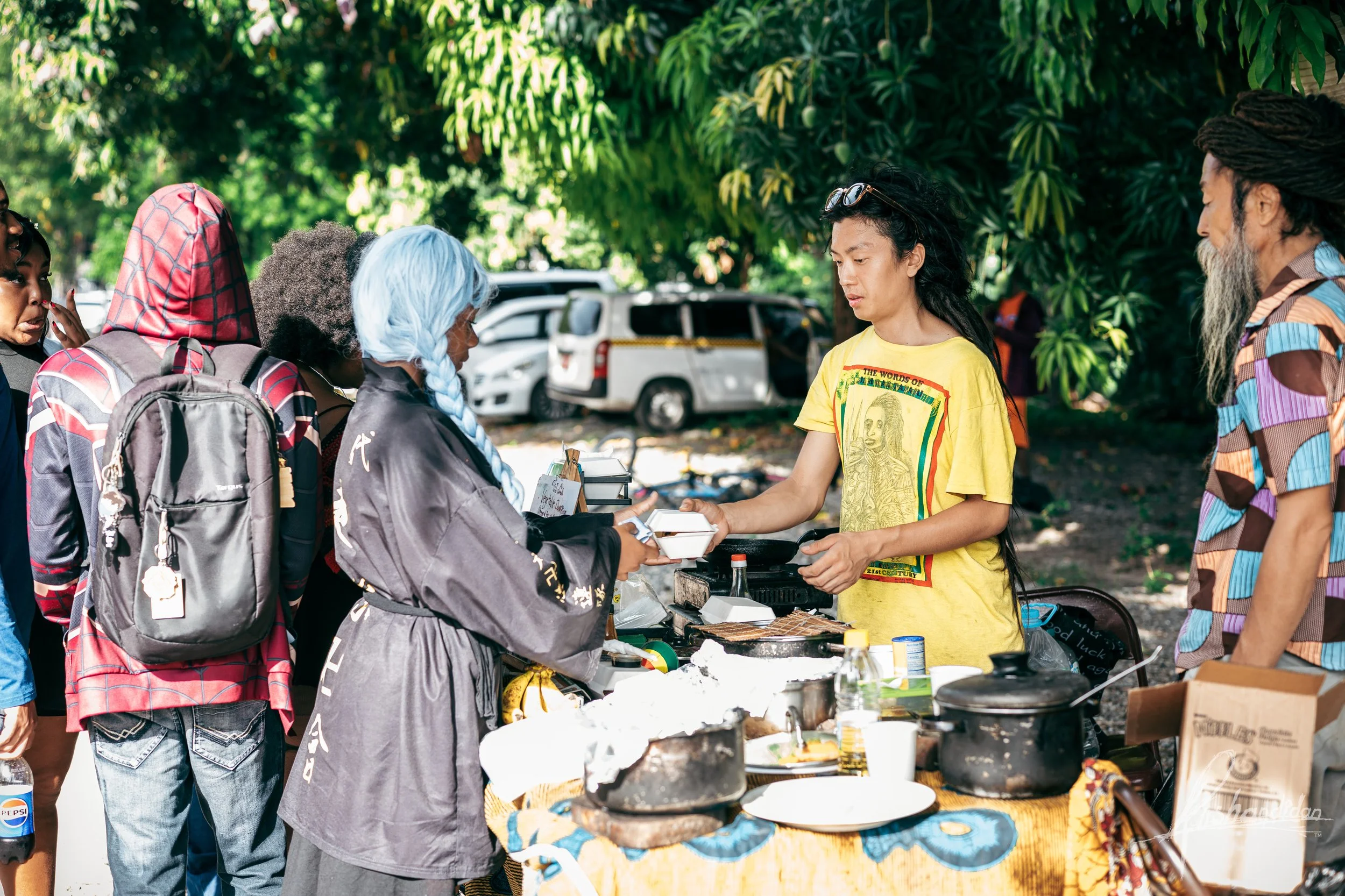 A group of people gathered at an outdoor food stand under green trees with parked cars in the background. A woman with long dark hair wearing a yellow T-shirt is serving food to customers.