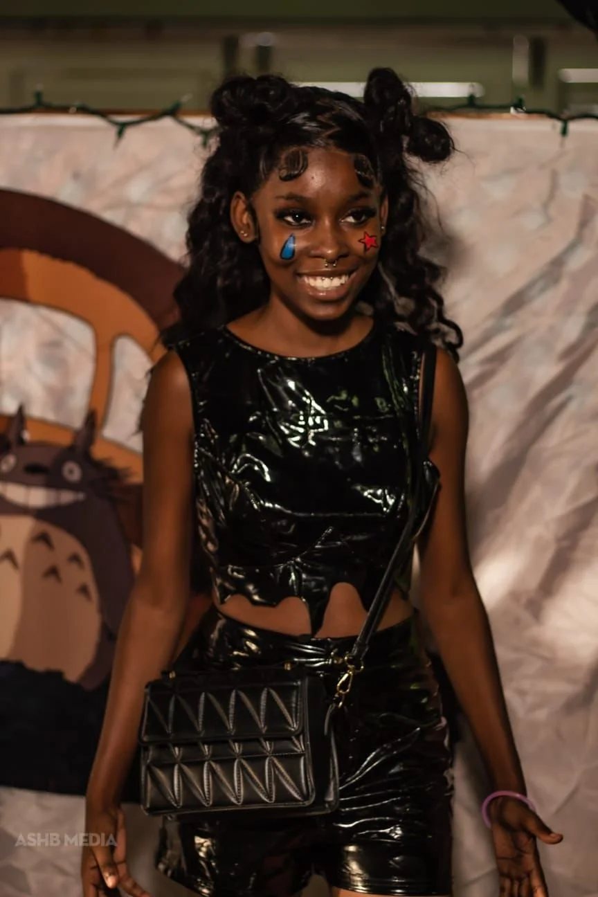 A young girl with dark curly hair styled in buns, smiling, wearing a shiny black sleeveless outfit, with face paint of a blue teardrop under her right eye and a red star under her left eye, holding a black quilted purse with a gold chain strap, stand