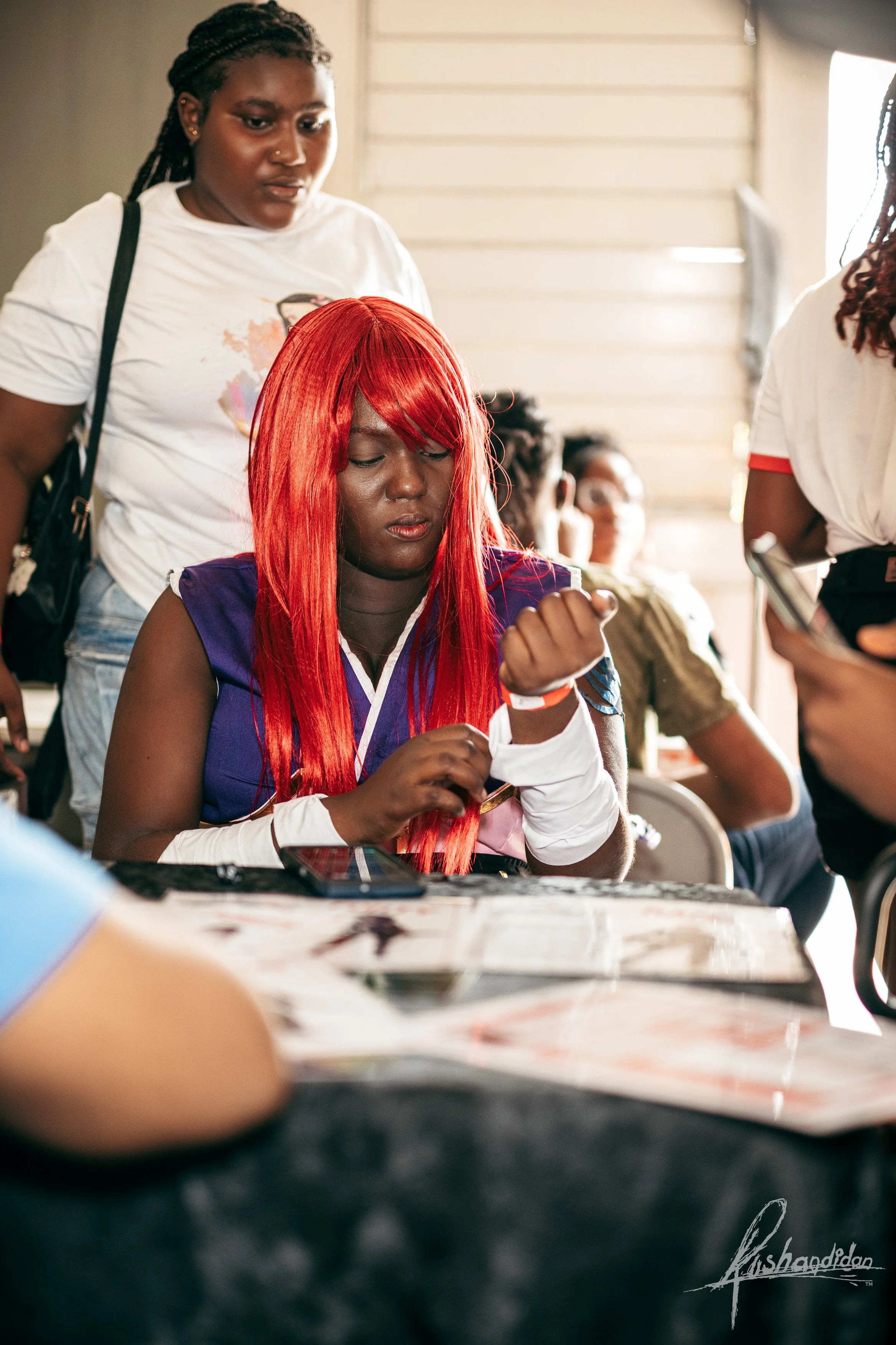 A woman with bright red hair is sitting at a table, looking down at her wrist, possibly adjusting a bracelet. Two women stand nearby, one behind her and one to her right. There are several people blurry in the background, and the setting appears to b