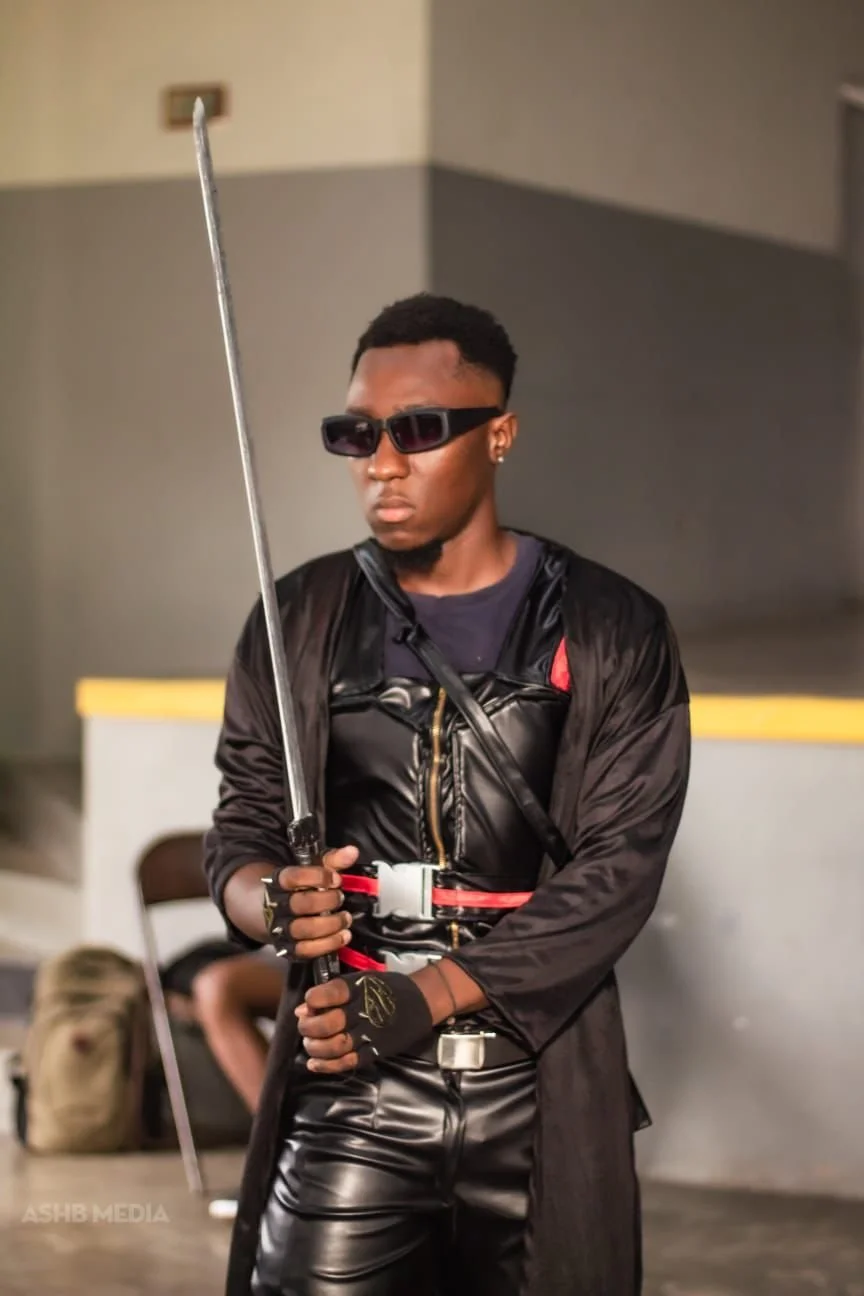 A young man dressed in black leather outfit, wearing sunglasses, holding a sword, standing in an indoor setting.