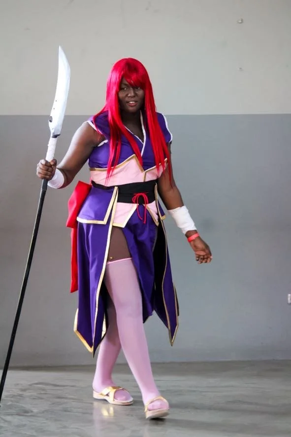 Person dressed in a colorful cosplay costume with a red wig, purple and pink outfit, thigh-high stockings, and holding a prop spear, standing indoors against a plain wall.