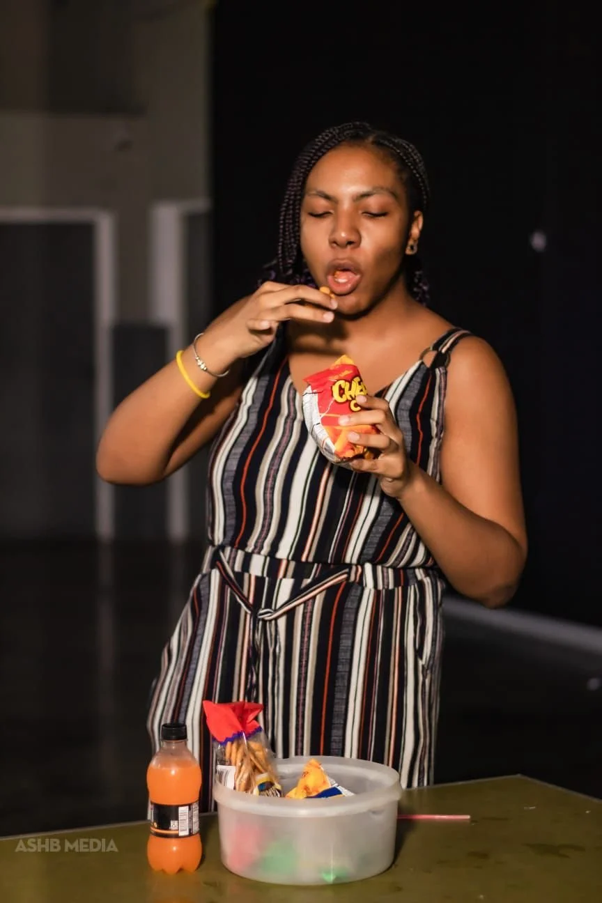 A woman wearing a striped jumpsuit is holding a bag of Cheetos and eating snacks. There is a bottle of orange drink and a container with snacks on the table in front of her.