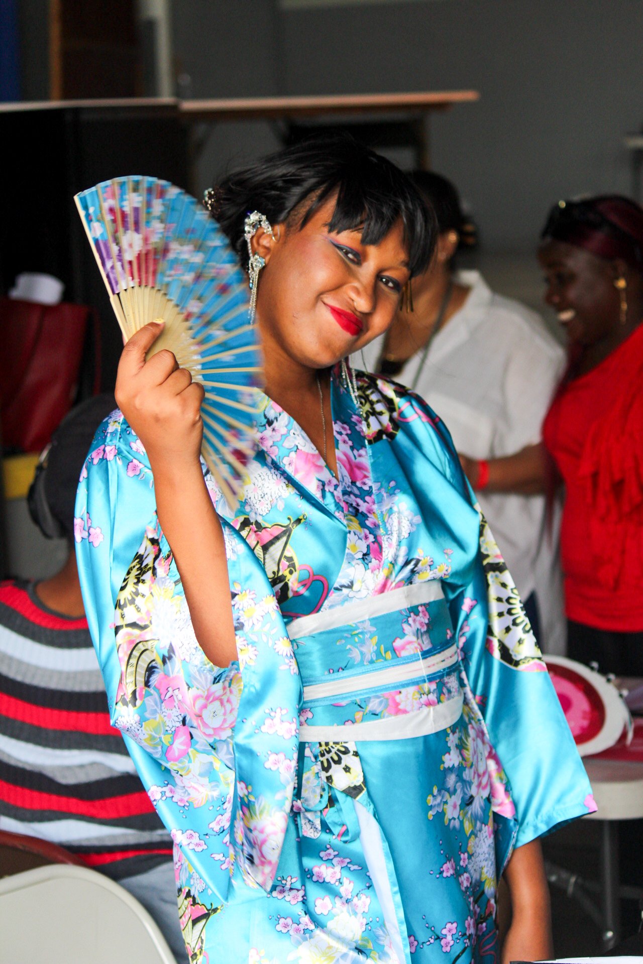 Woman in a blue floral kimono holding a colorful paper fan, smiling, with other people in the background.