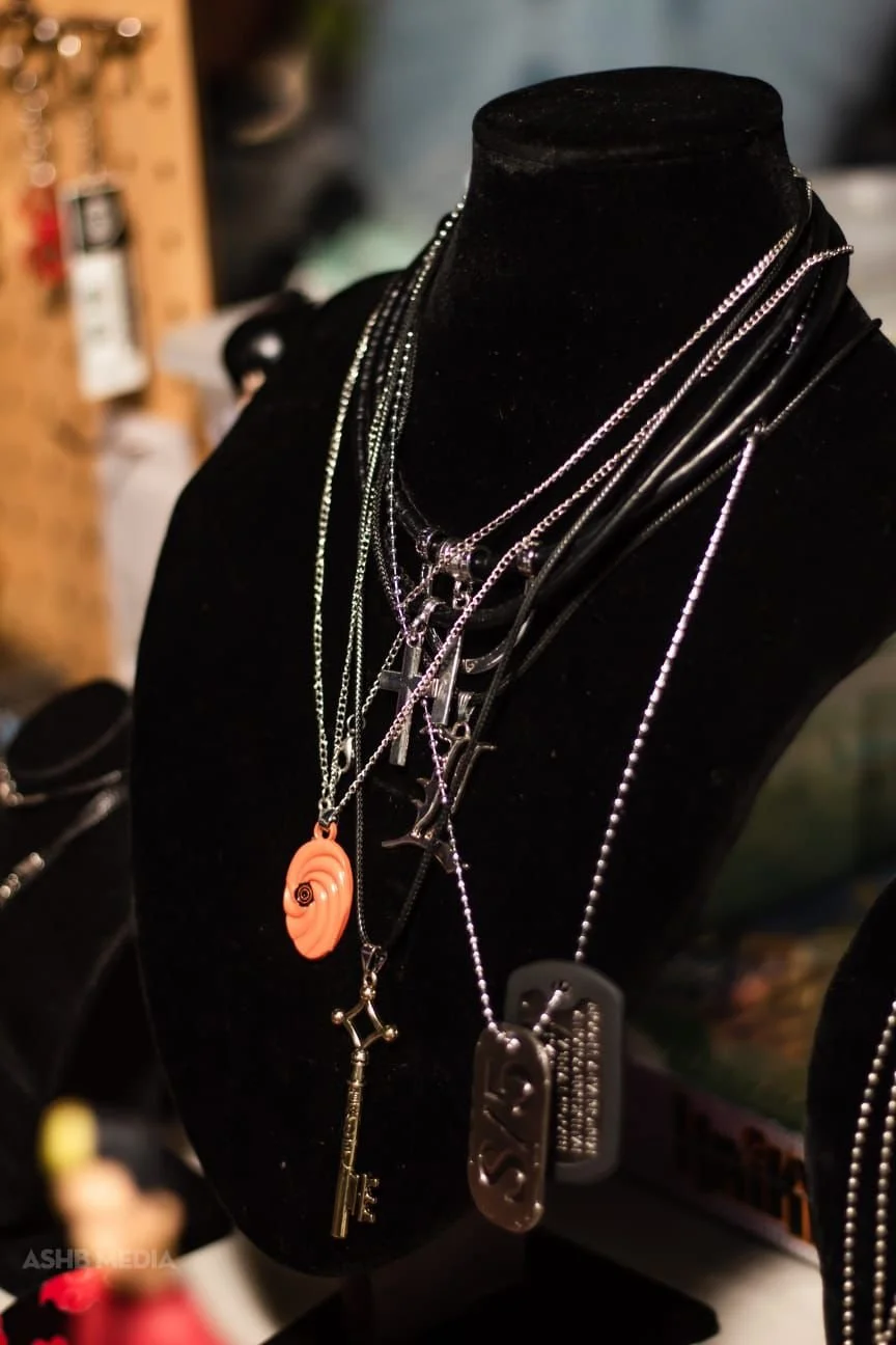 A black jewelry display bust showcasing multiple silver, black, and gold layered necklaces, with pendants including a shell, a key, a dog tag, and various charms.