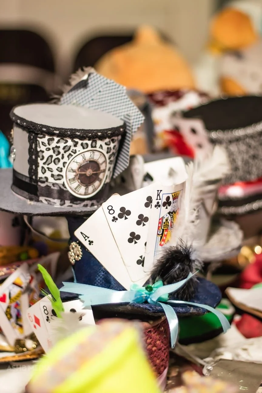A collection of small decorative hats and a fan of playing cards, including an ace of spades, eight of clubs, king of clubs, and queen of hearts, arranged on a surface.