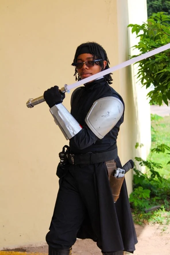 Person dressed in black with armor on shoulder, holding a white lightsaber, standing beside a yellow wall and green plants.