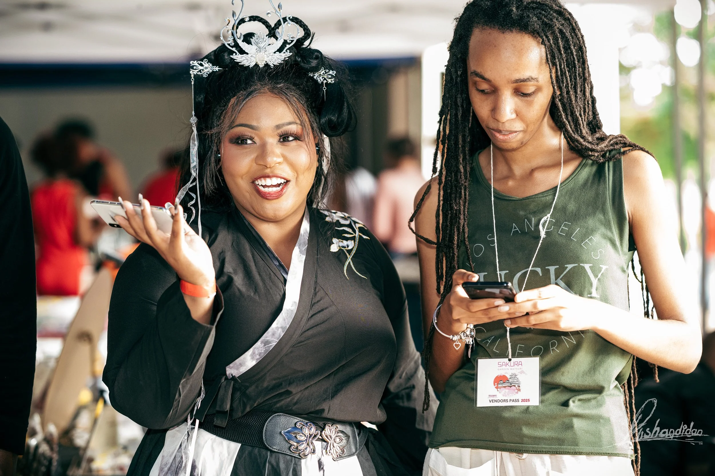 Two women, one dressed as a character with a crown and formal attire, smiling and talking, and the other in casual green tank top with a vendor pass, looking at her phone at an event or convention.