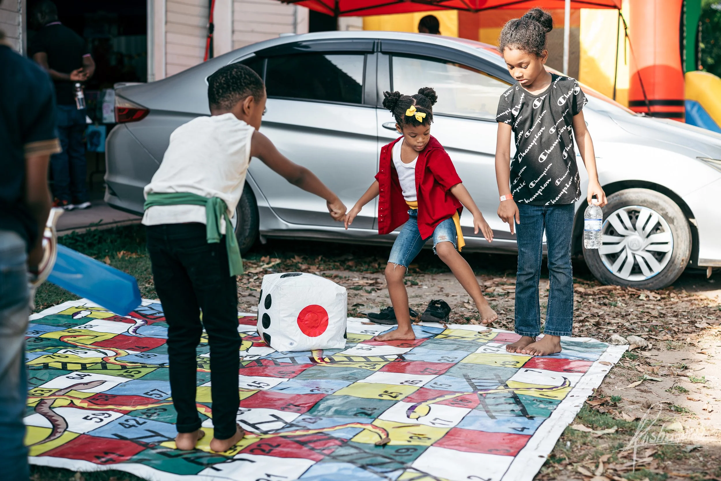 Children playing a game of Twister in an outdoor area with a large die nearby, under a canopy next to a silver car.