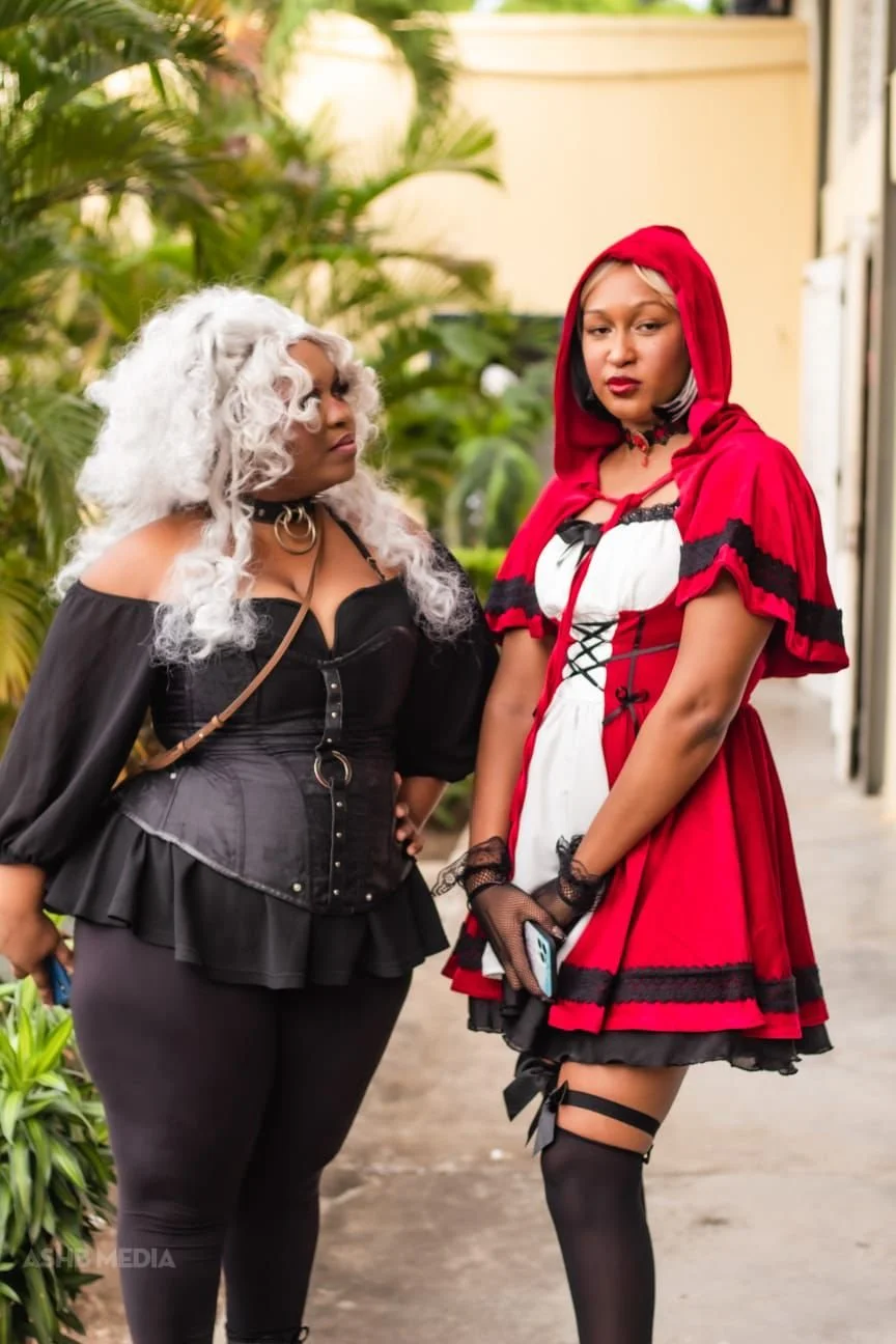 Two women dressed in cosplay costumes standing outdoors surrounded by greenery. One woman is in a black gothic-inspired outfit with a white curly wig, and the other woman is in a red and white costume with a hood and black stockings.