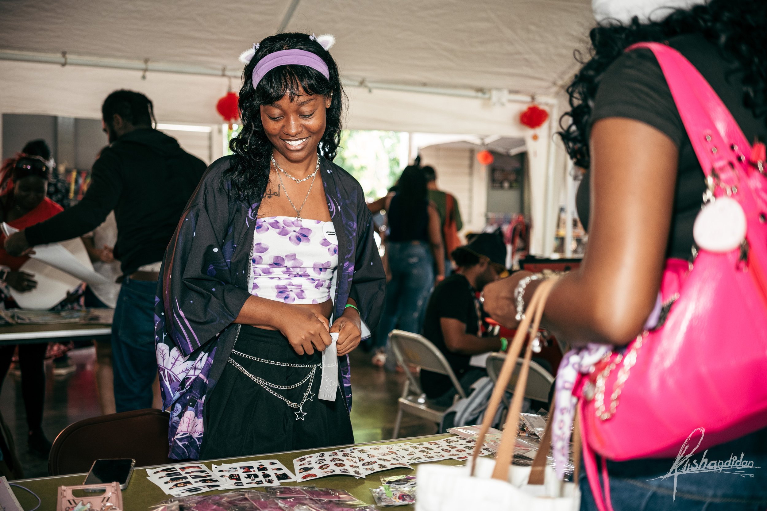 A young woman with dark curly hair, purple headband, and jewelry shopping at a market stall with jewelry and photos on display, smiling as she looks at the items.