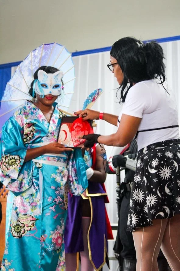 A person dressed in a colorful kimono and wearing a fox mask holding a red and black box, standing next to a woman in a white top and starry skirt, who is pointing at the box. There are other people in costumes in the background.