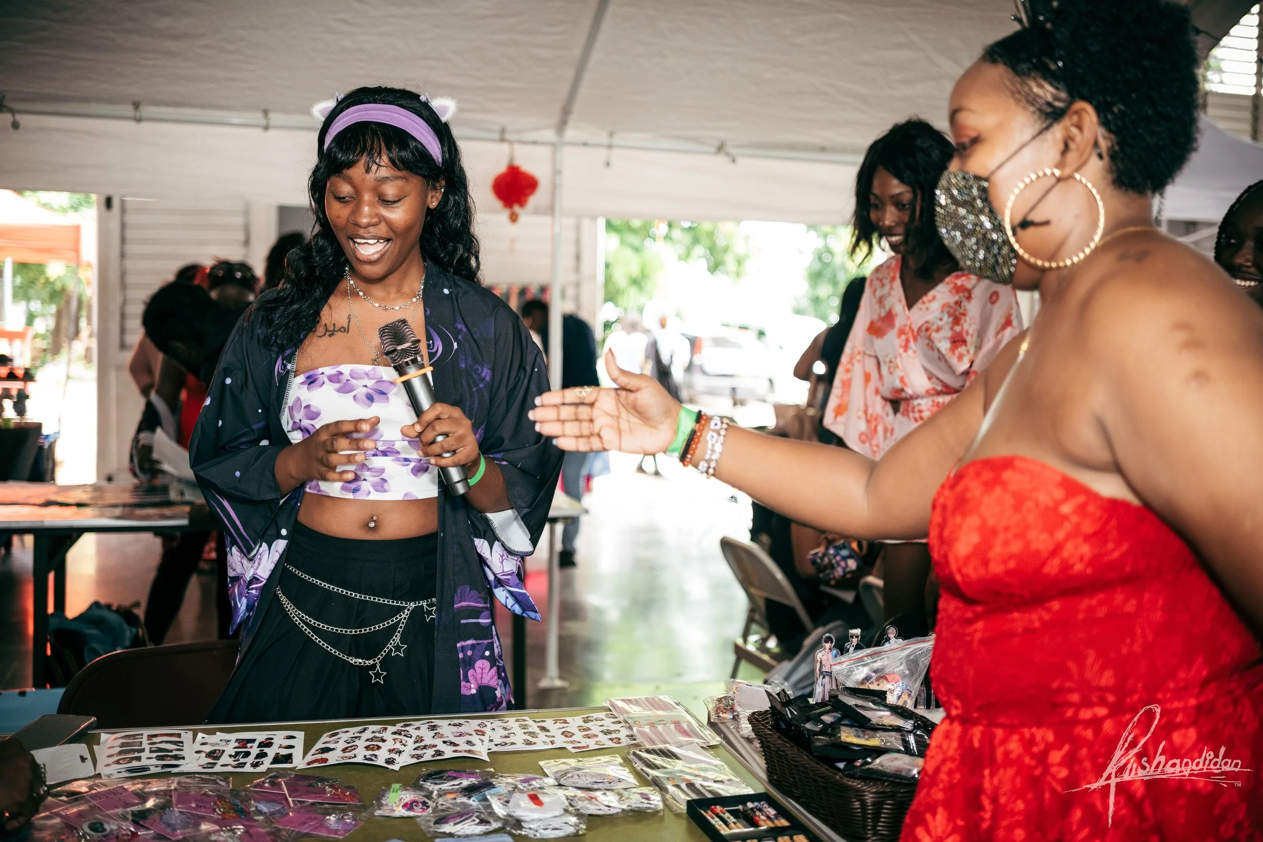 A young woman at a market stall holding a microphone, smiling and engaging with a woman wearing a red dress and face mask. Other shoppers are in the background under a white tent.