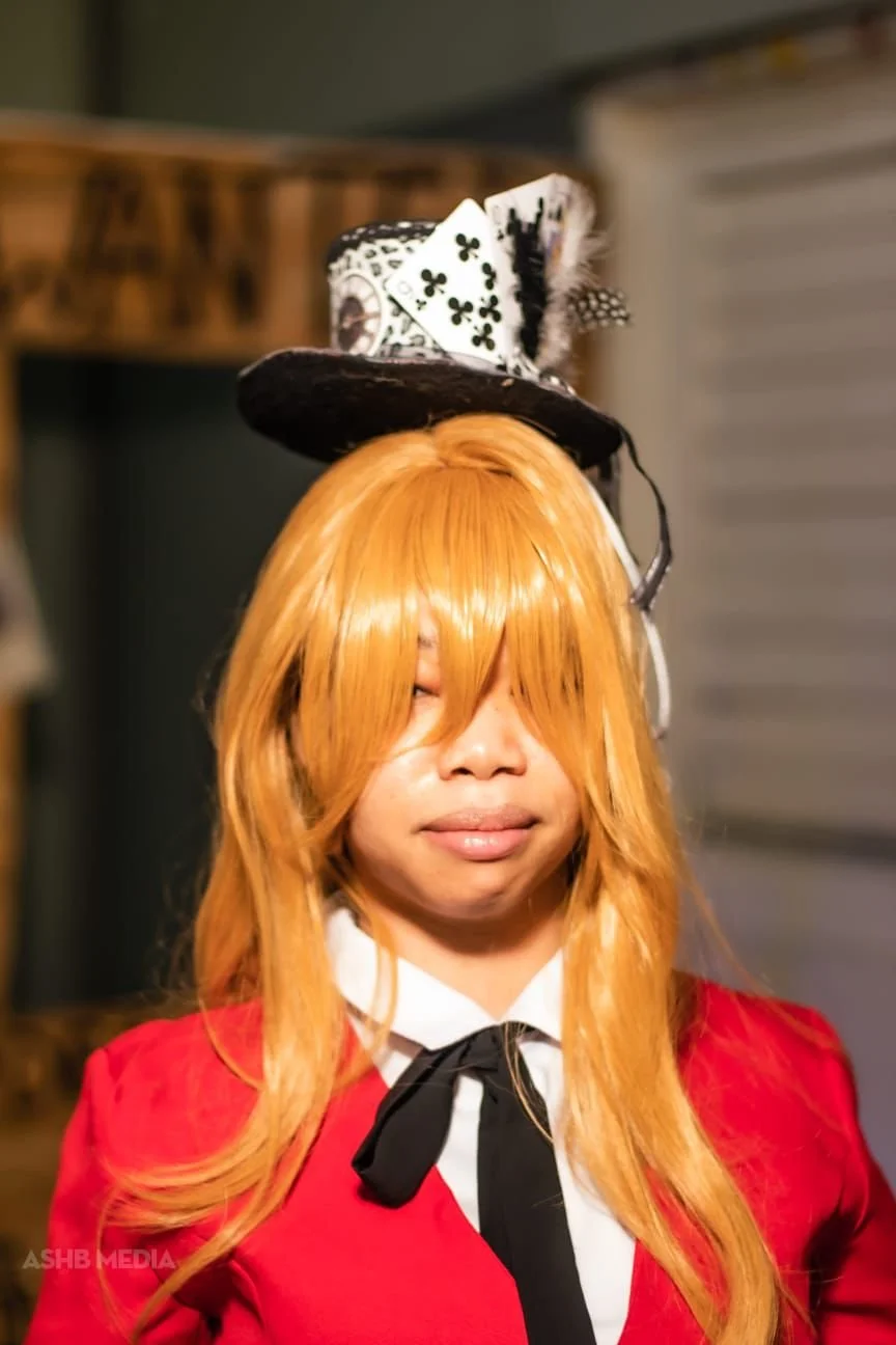 Person with long orange hair wearing a red jacket, white shirt with black tie, and a small hat with playing cards on top.