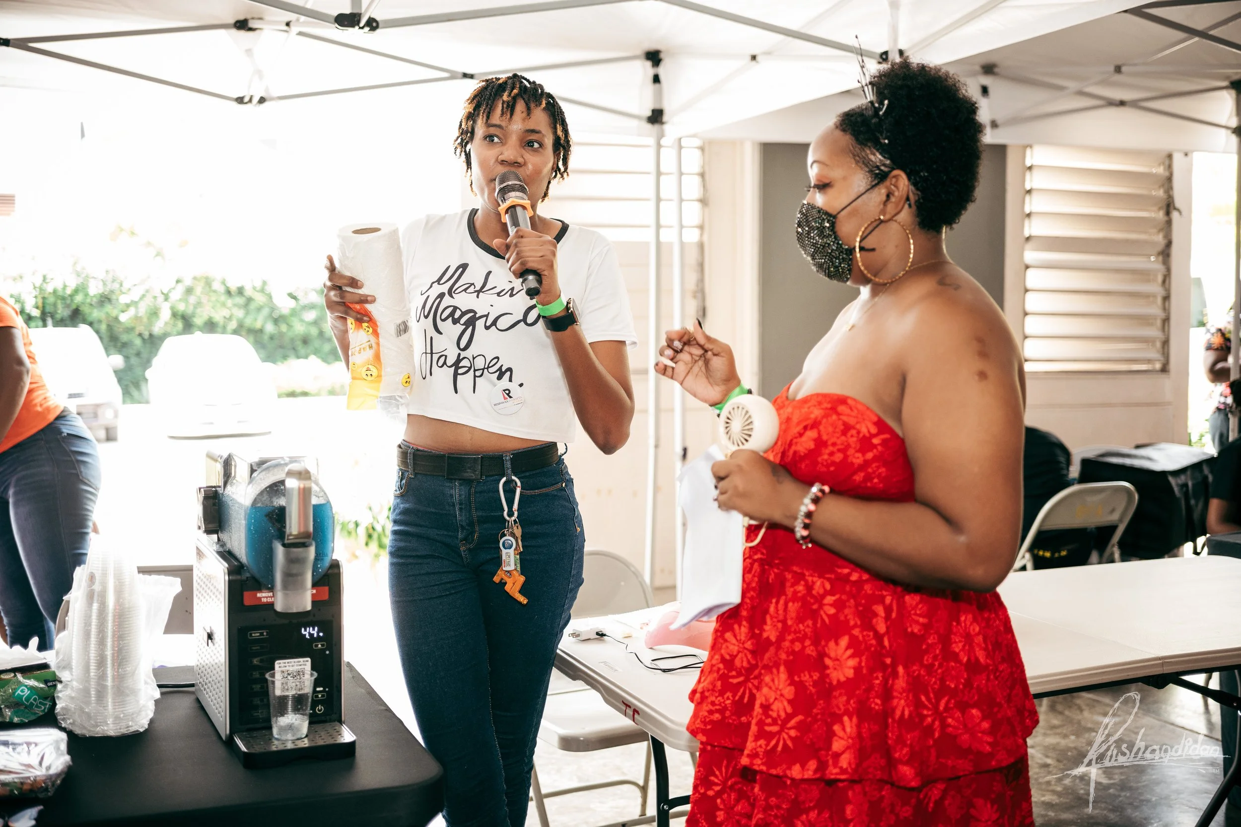 Two women standing under a white canopy tent, one speaking into a microphone while holding a roll of paper towels, the other wearing a face mask and holding a small handheld fan. The woman with the microphone is wearing a white crop top with text, an