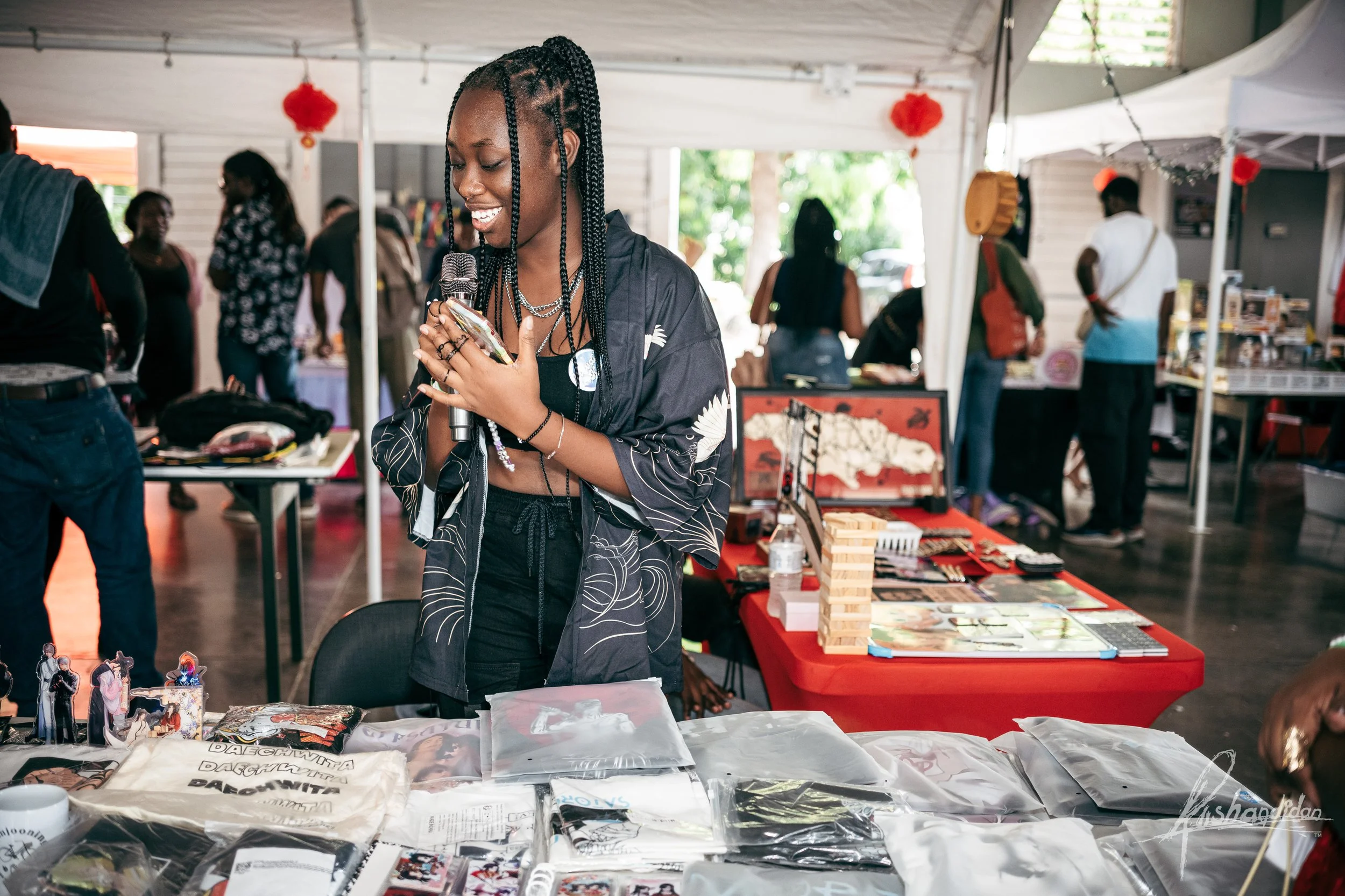 A woman with braided hair smiling and holding a microphone at a booth with various merchandise, including T-shirts, art prints, and accessories, in a lively indoor market setting.