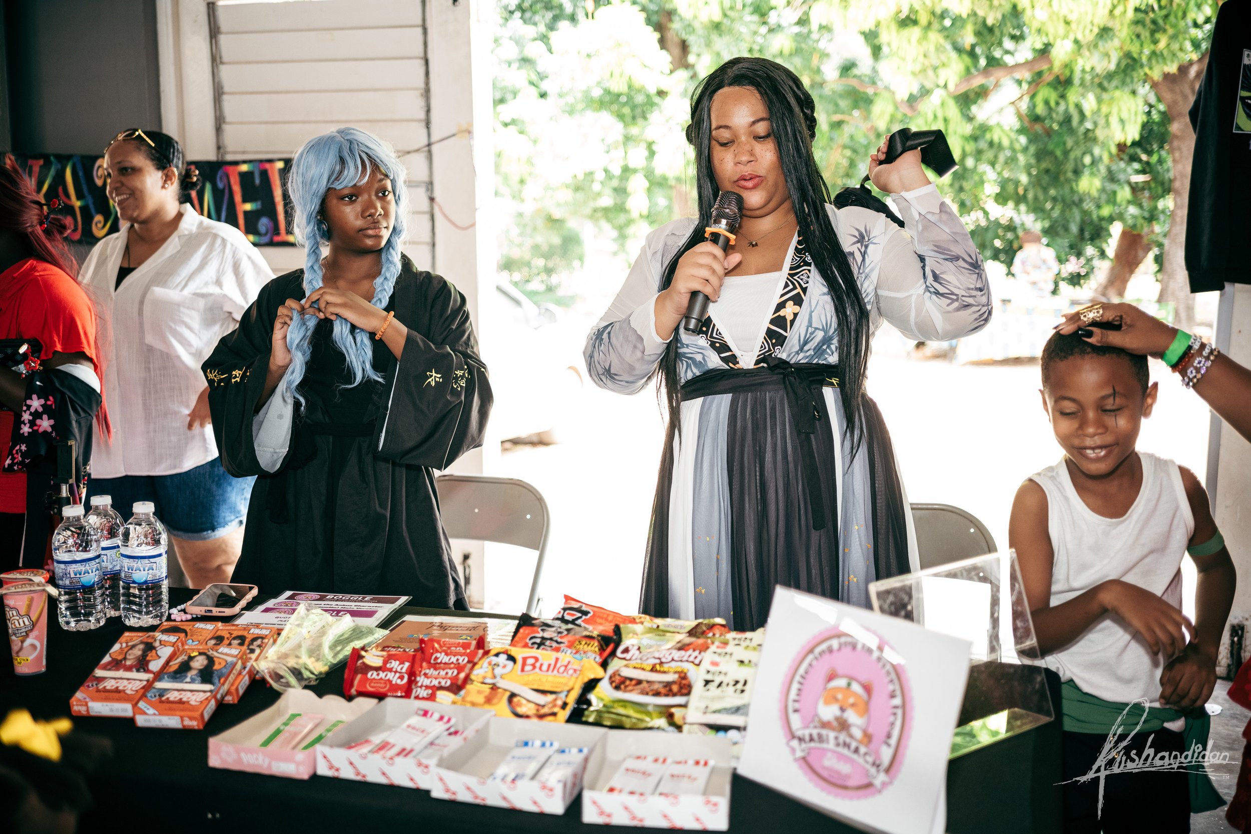 A group of young people at a snack booth during a celebration. A woman with long black hair is holding a microphone and appears to be speaking or singing. A girl with blue hair is standing nearby, and another young girl with short hair is smiling as 