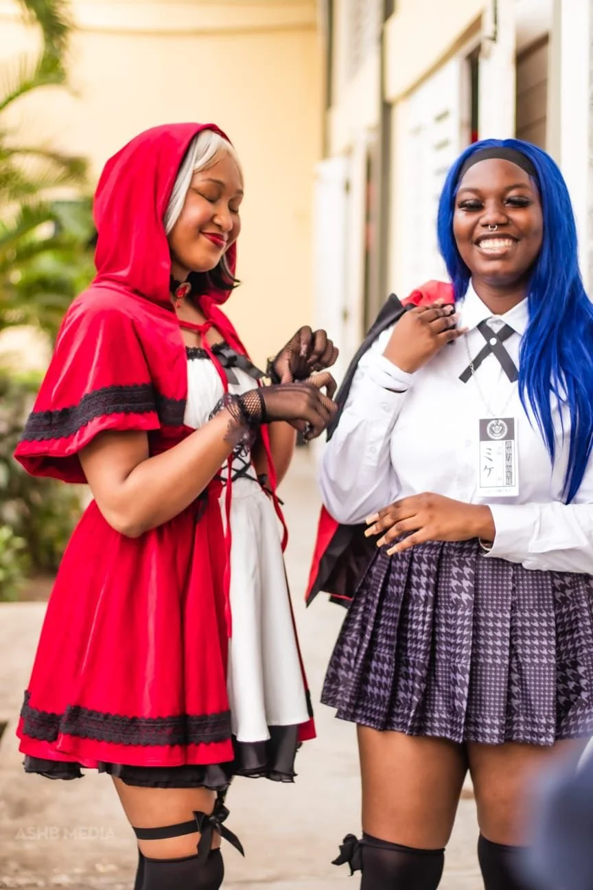 Two women smiling outdoors, one dressed as Little Red Riding Hood and the other in a school uniform with bright blue hair.