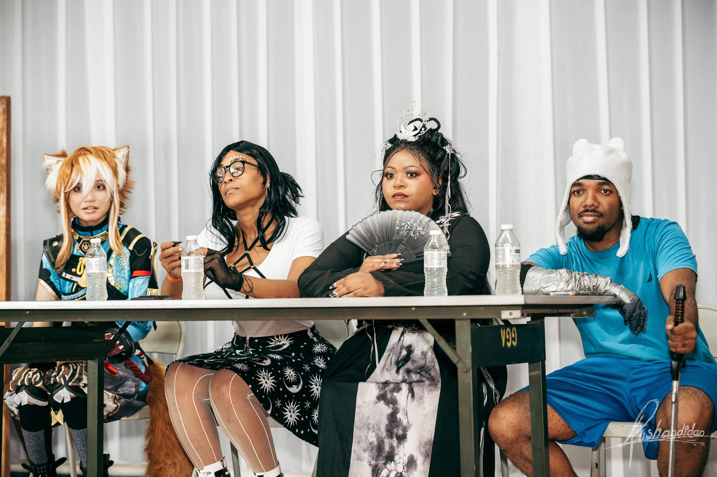 Four people sitting at a table dressed in cosplay costumes, with water bottles in front of them, at a convention or event.