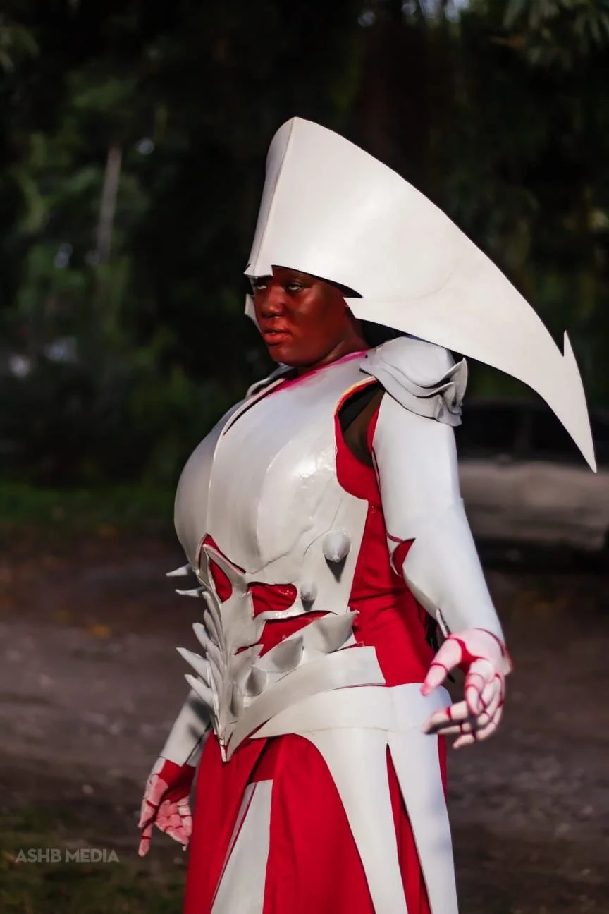 A person dressed in elaborate fantasy armor with a large white headpiece and red and white attire, standing outdoors with trees in the background.