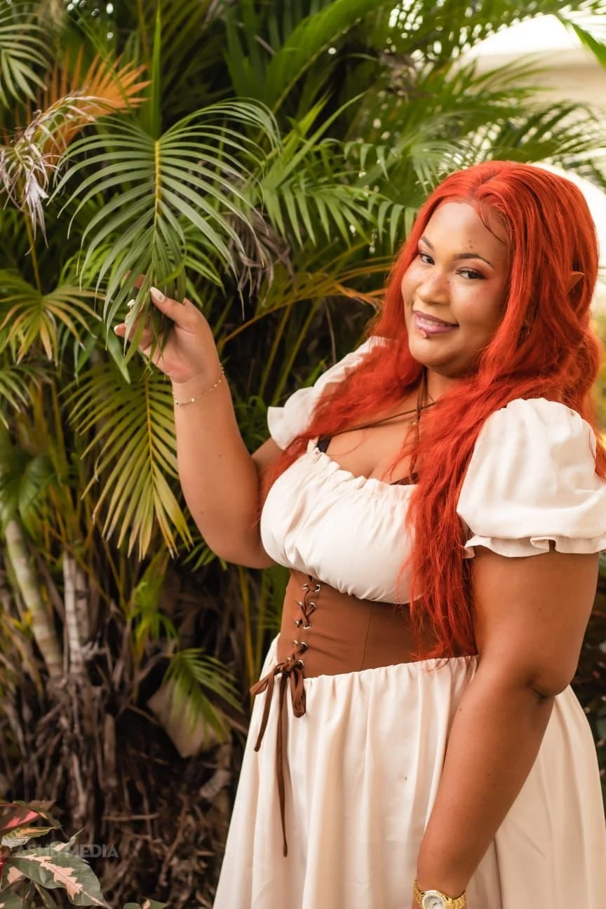 A woman with long red hair smiling and holding a palm leaf, standing in front of lush green tropical plants.
