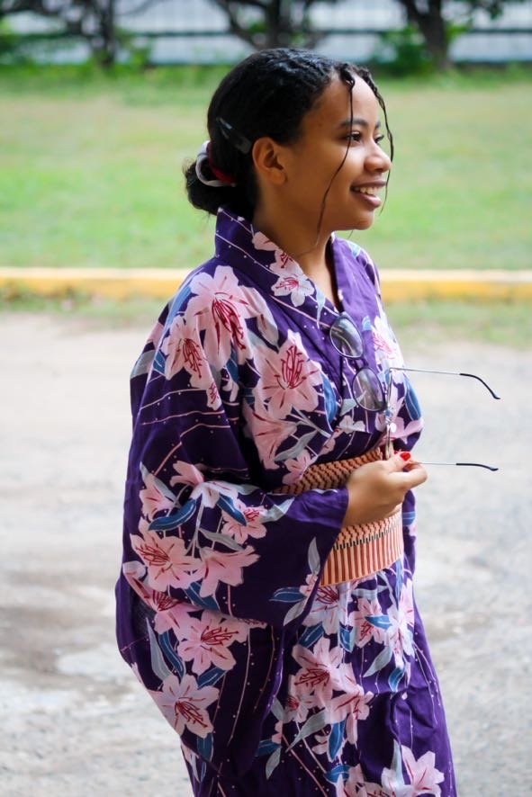A woman in a purple floral yukata holding glasses, smiling while standing outdoors.