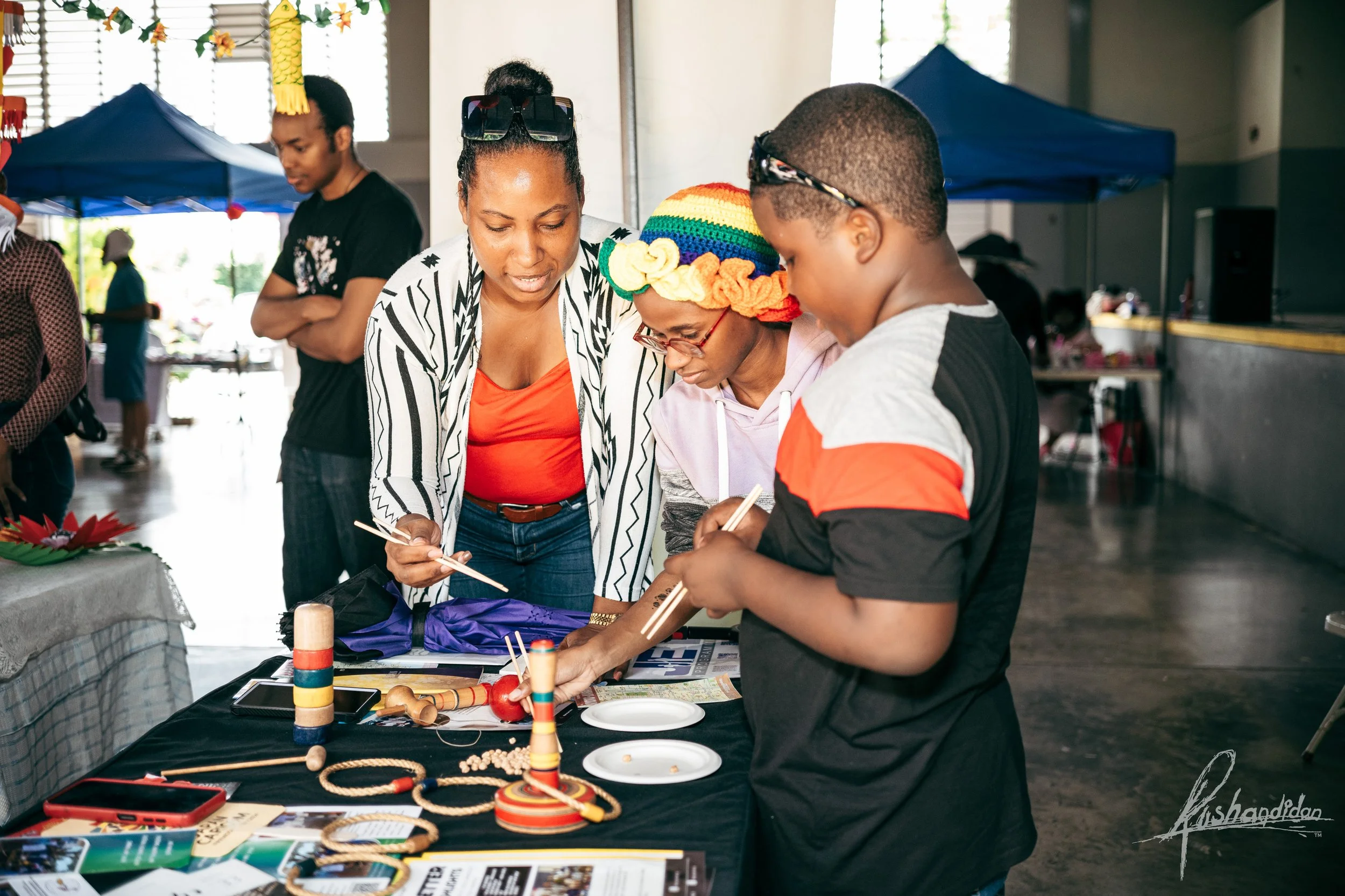 Three people examining handmade jewelry at a booth with craft items, including necklaces and bracelets, during an indoor event.