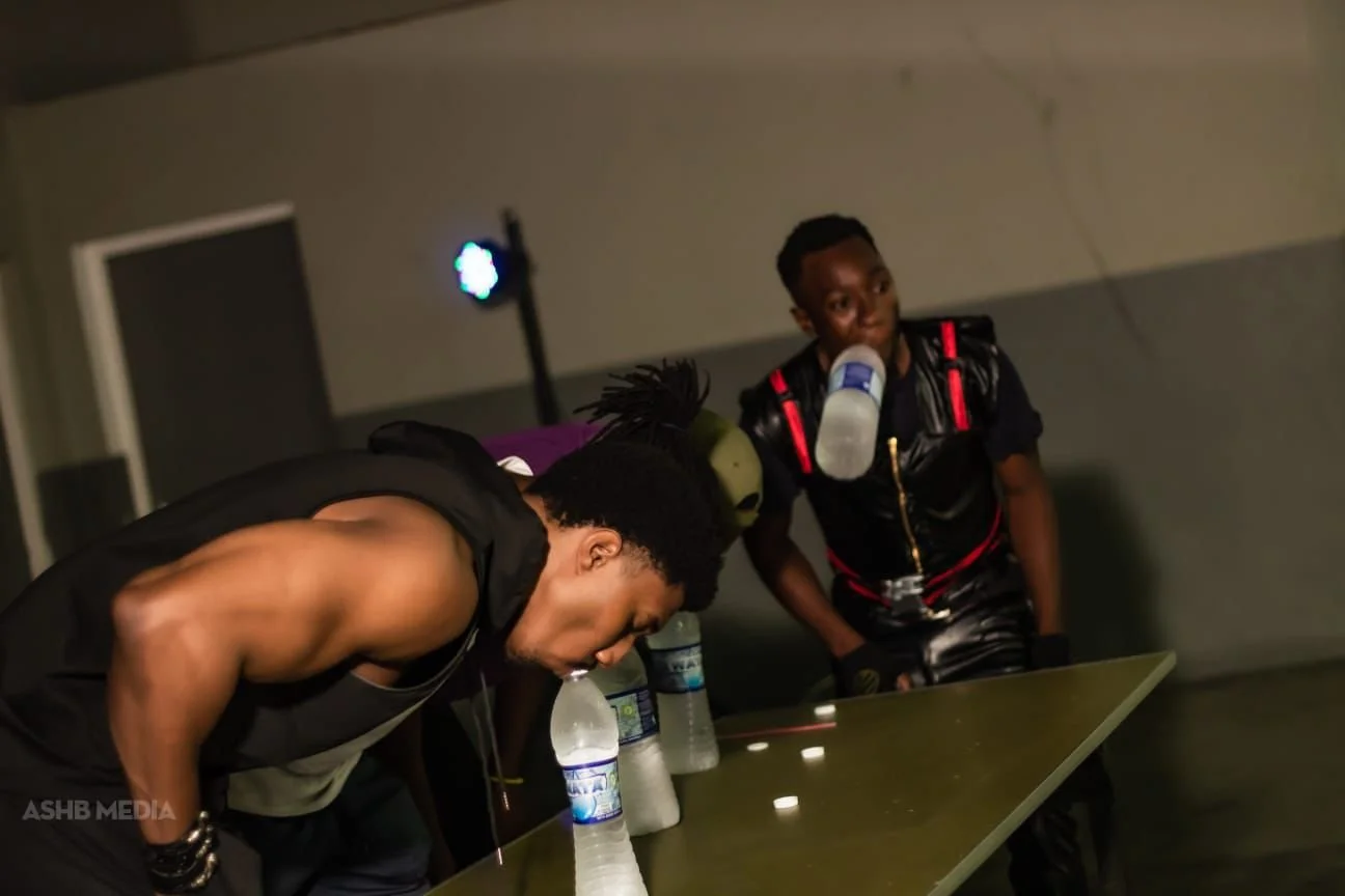 Two young men are at a table with bottles of water. One is bending over with his head down, and the other is standing, drinking from a water bottle, with a small stage light and a plain wall in the background.