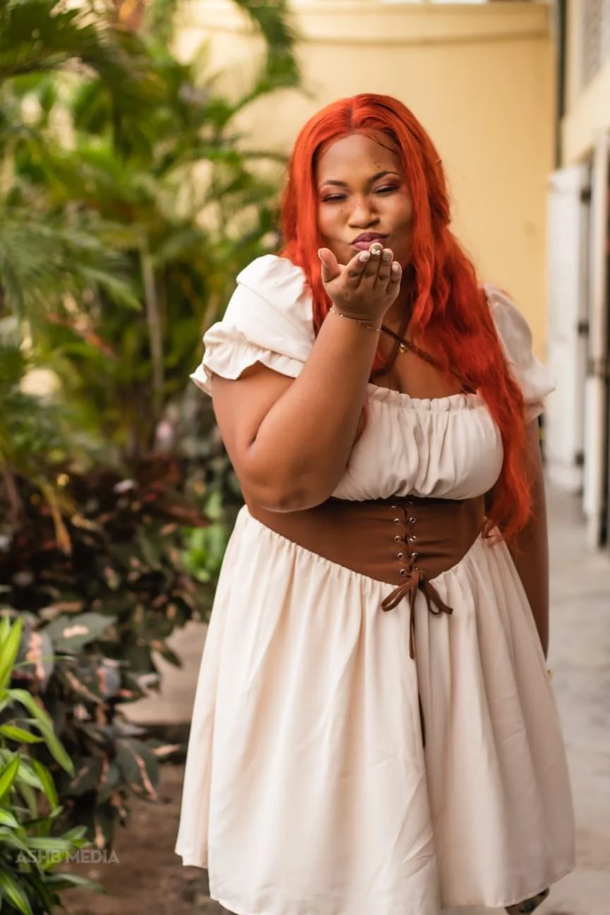 A woman with long red hair blowing a kiss in an outdoor setting with greenery in the background.
