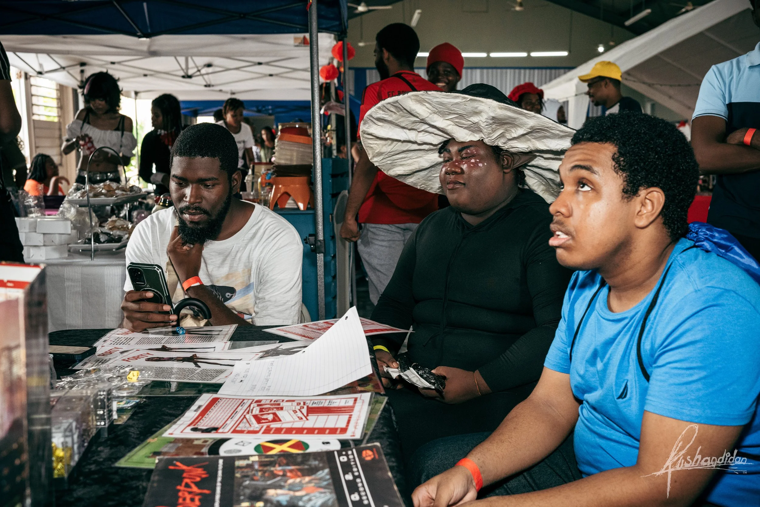 Three young men sitting at a table at a market or fair, with various game and event materials spread out in front of them. The man on the left is looking at his phone, the woman in the middle is wearing a large, crinkled hat, and the man on the right
