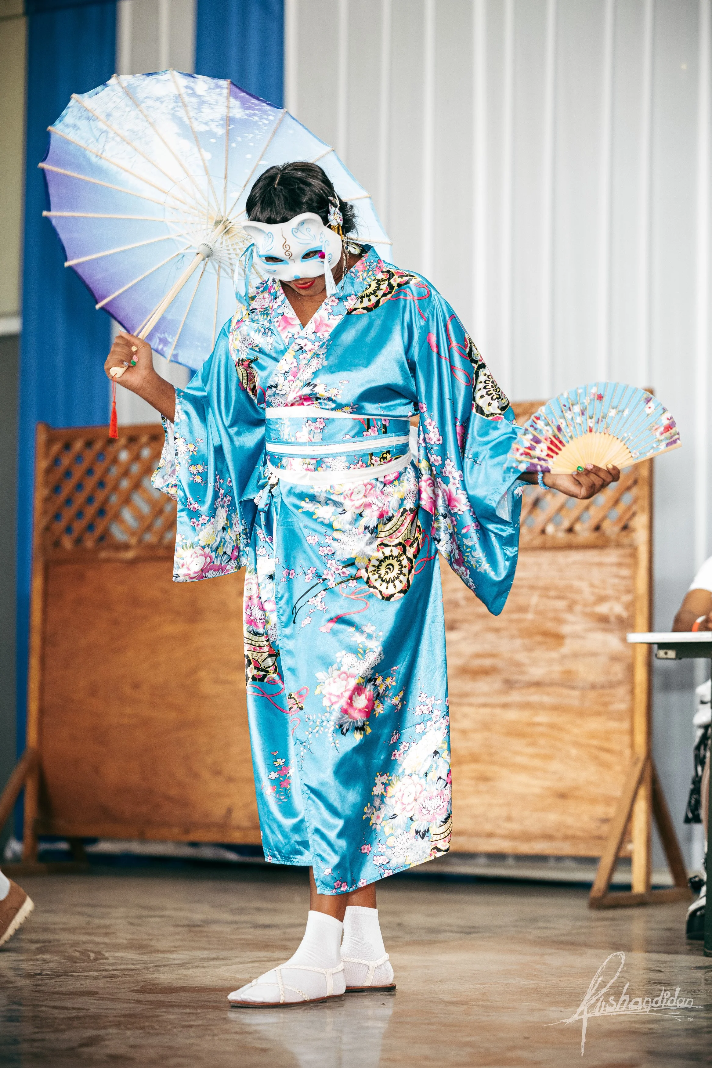 Person dressed in a vibrant blue kimono with floral and traditional patterns, wearing a white mask, holding a paper parasol and a fan, performing a dance.
