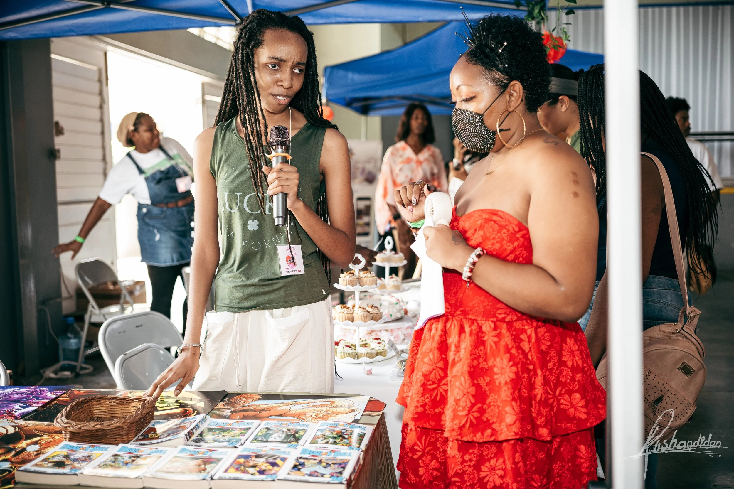 Women at an outdoor market booth discussing. One woman holds a microphone, another a small hand-held fan. Market table has magazines, jewelry, and baked goods, including cupcakes.