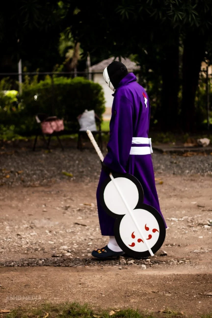 Person dressed as a traditional Japanese snow woman with a purple coat, black hood, and white face mask walking outdoors on a dirt path, carrying a large, stylized shamisen with a face design.