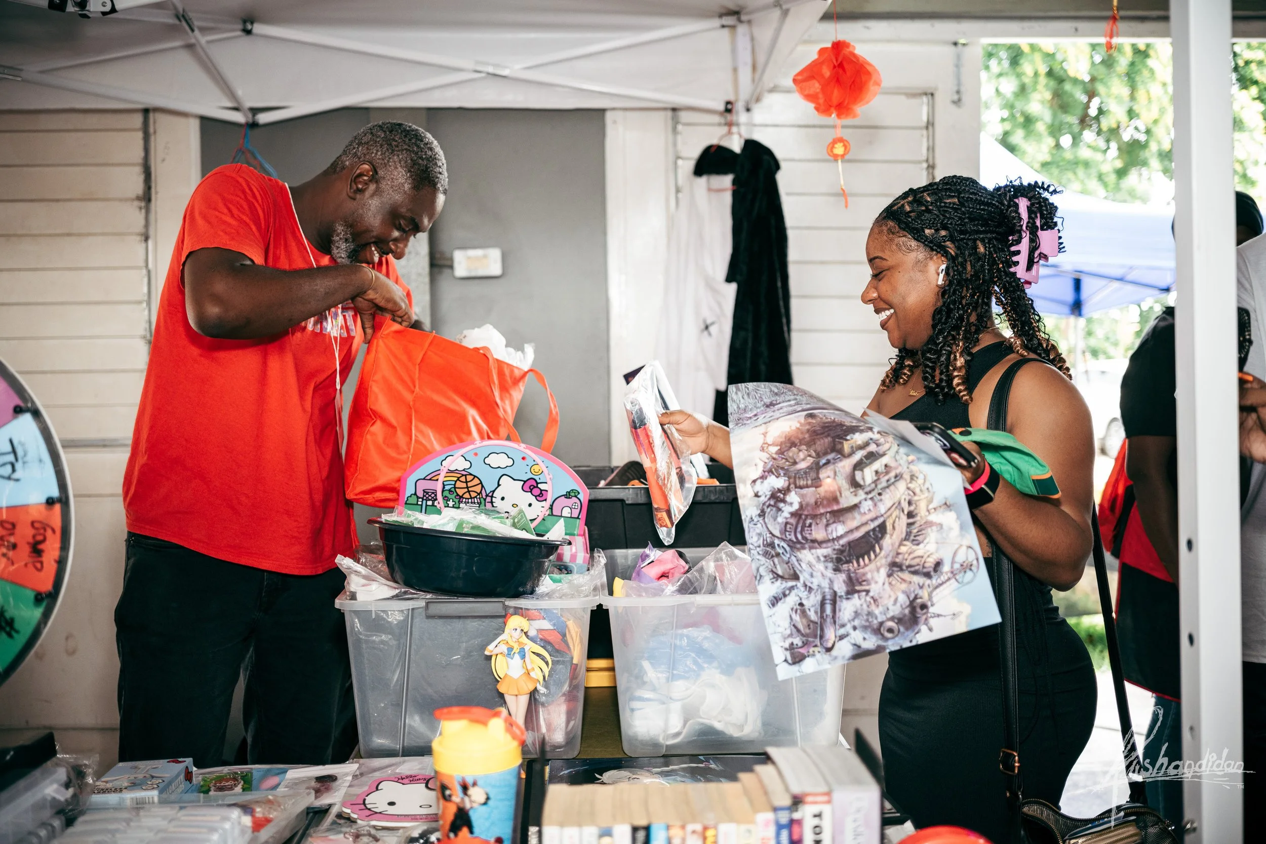 A man in a red shirt and a woman in a black top smiling at each other at an outdoor market stall selling toys and collectibles, with various items and a spinning wheel in front of them.
