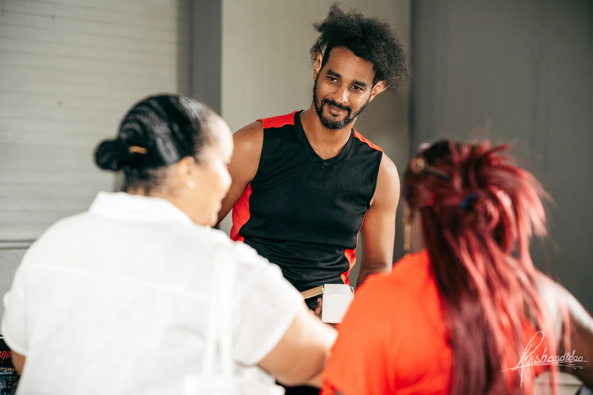 A man with curly hair and a beard wearing a sleeveless black athletic shirt with red accents, smiling and looking at two women seated in front of him. One woman has dark hair tied back and wears a white shirt, and the other has bright red hair and we