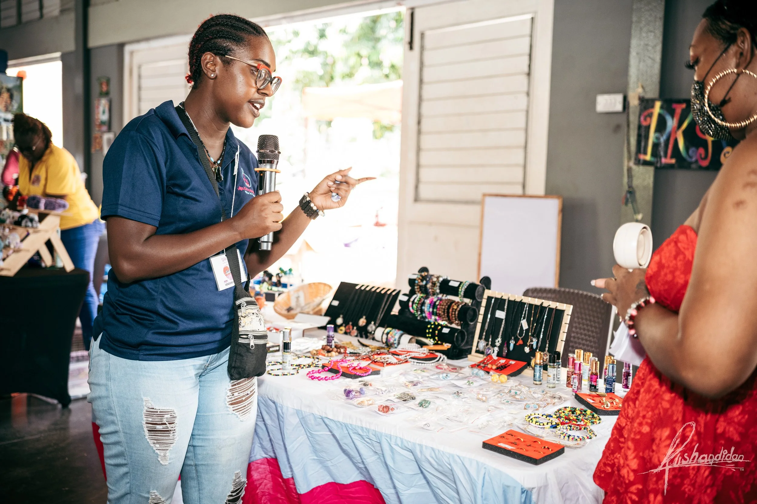 A woman with glasses and a navy blue shirt is talking and pointing at a jewelry display table. The table has various jewelry items including necklaces, bracelets, and earrings. Another woman in a red dress is browsing the jewelry, holding some pieces